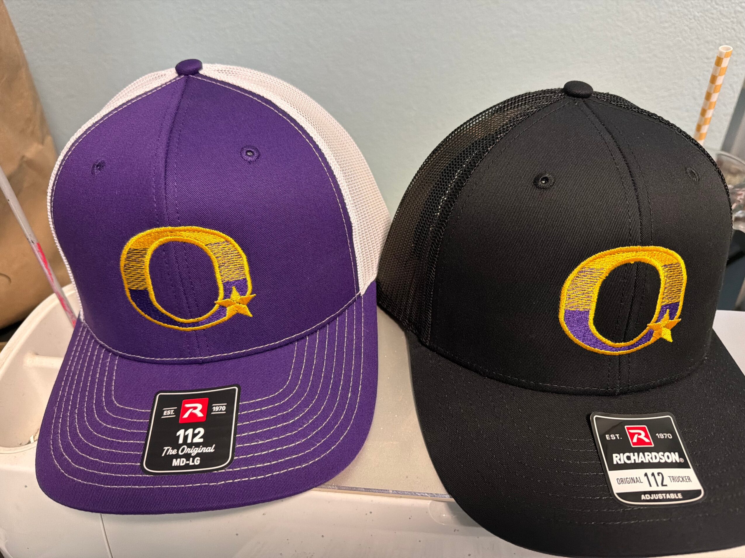 Two baseball caps, one purple and white, the other black, with a yellow and purple logo, placed on a white surface.