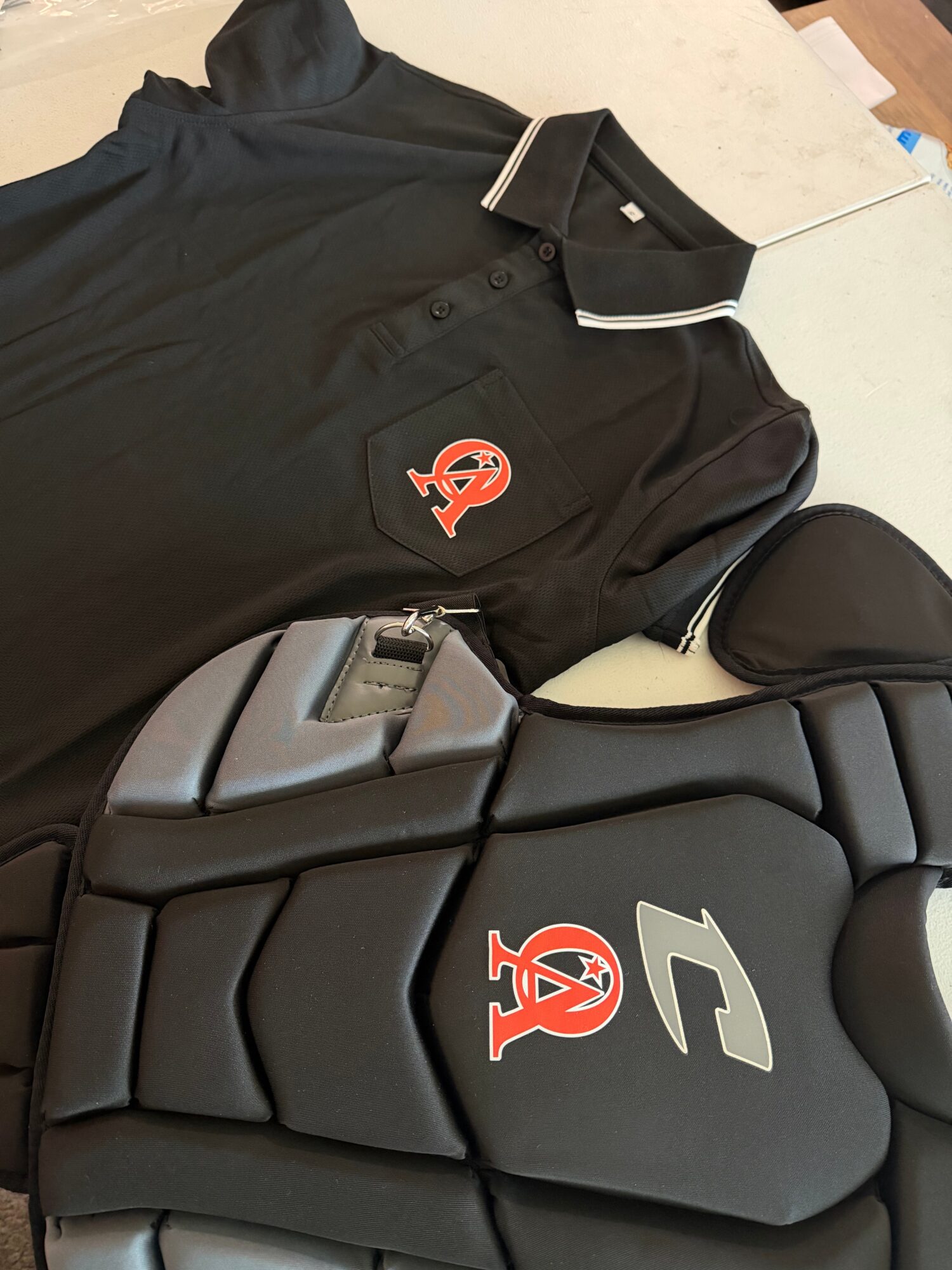 Black polo shirt with a logo, a black cap, and a black padded object on a white surface.