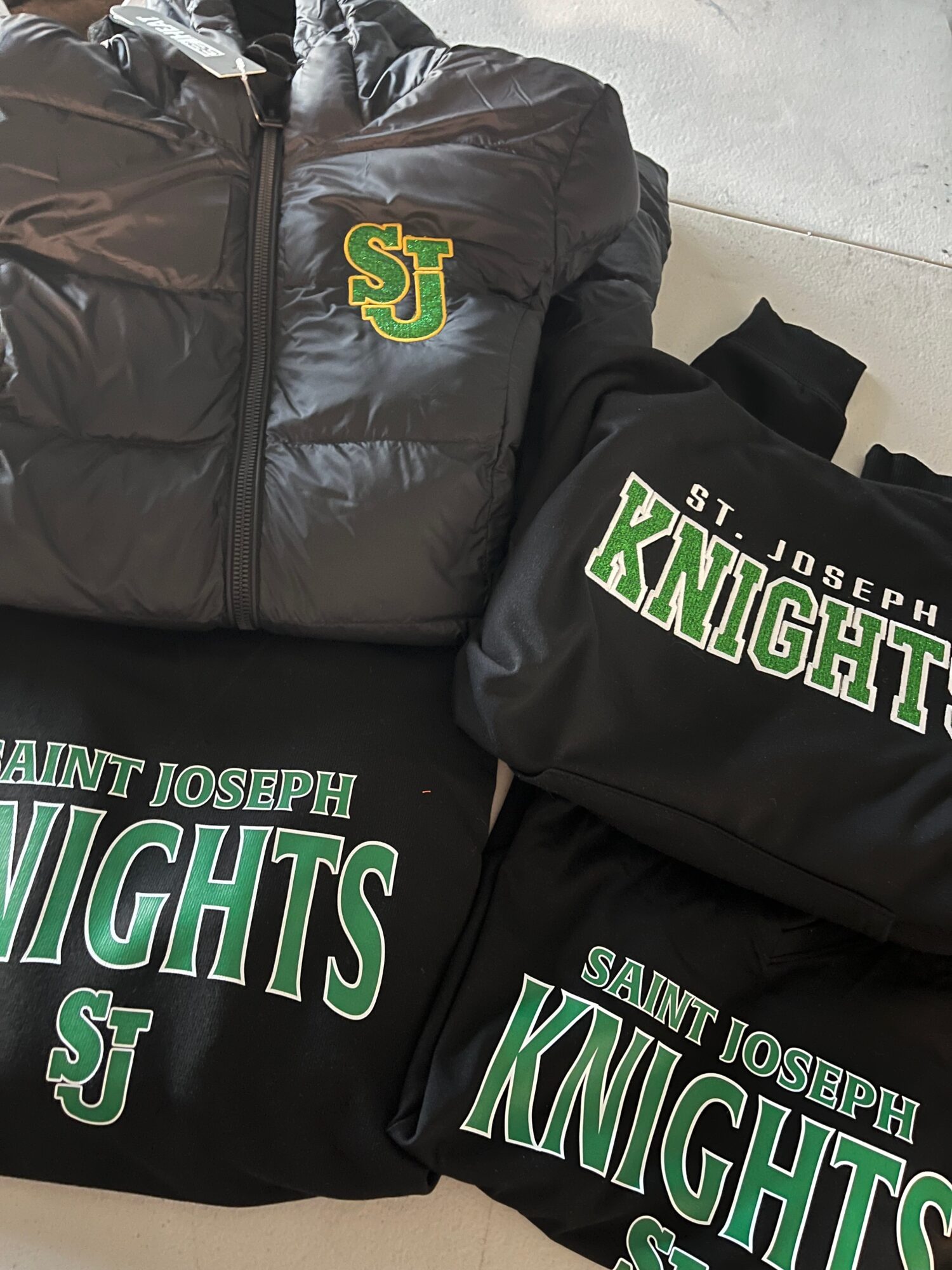 Black jacket, bag, and clothing items with Saint Joseph Knights logos and text, displayed on a surface.