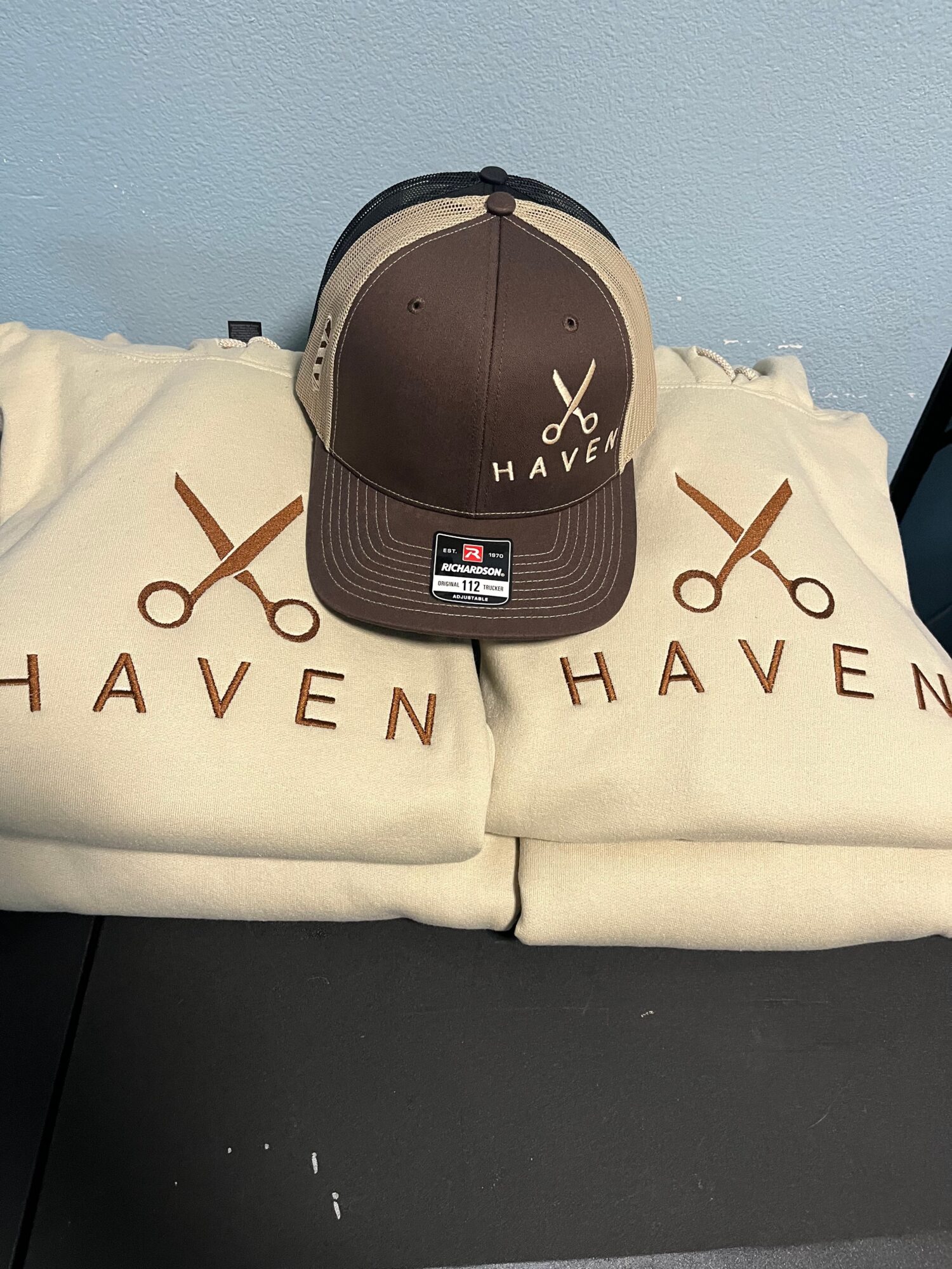Cap with scissors and 'HAVE' text, placed on two cloth bags with similar design, against a light blue wall.