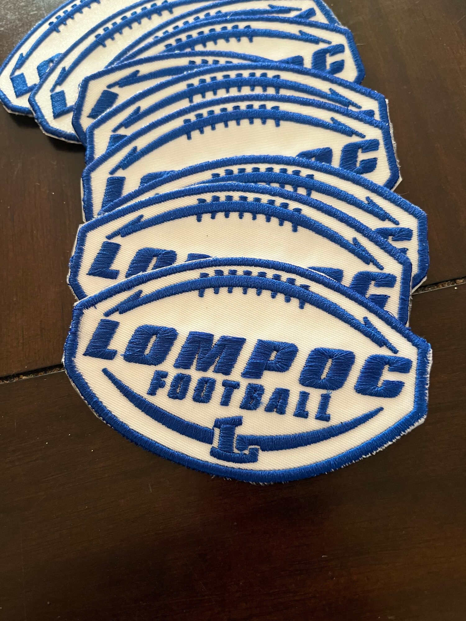 Stack of embroidered patches with 'LOMPOC FOOTBALL' text and a football shape, blue and white colors, on a wooden surface.