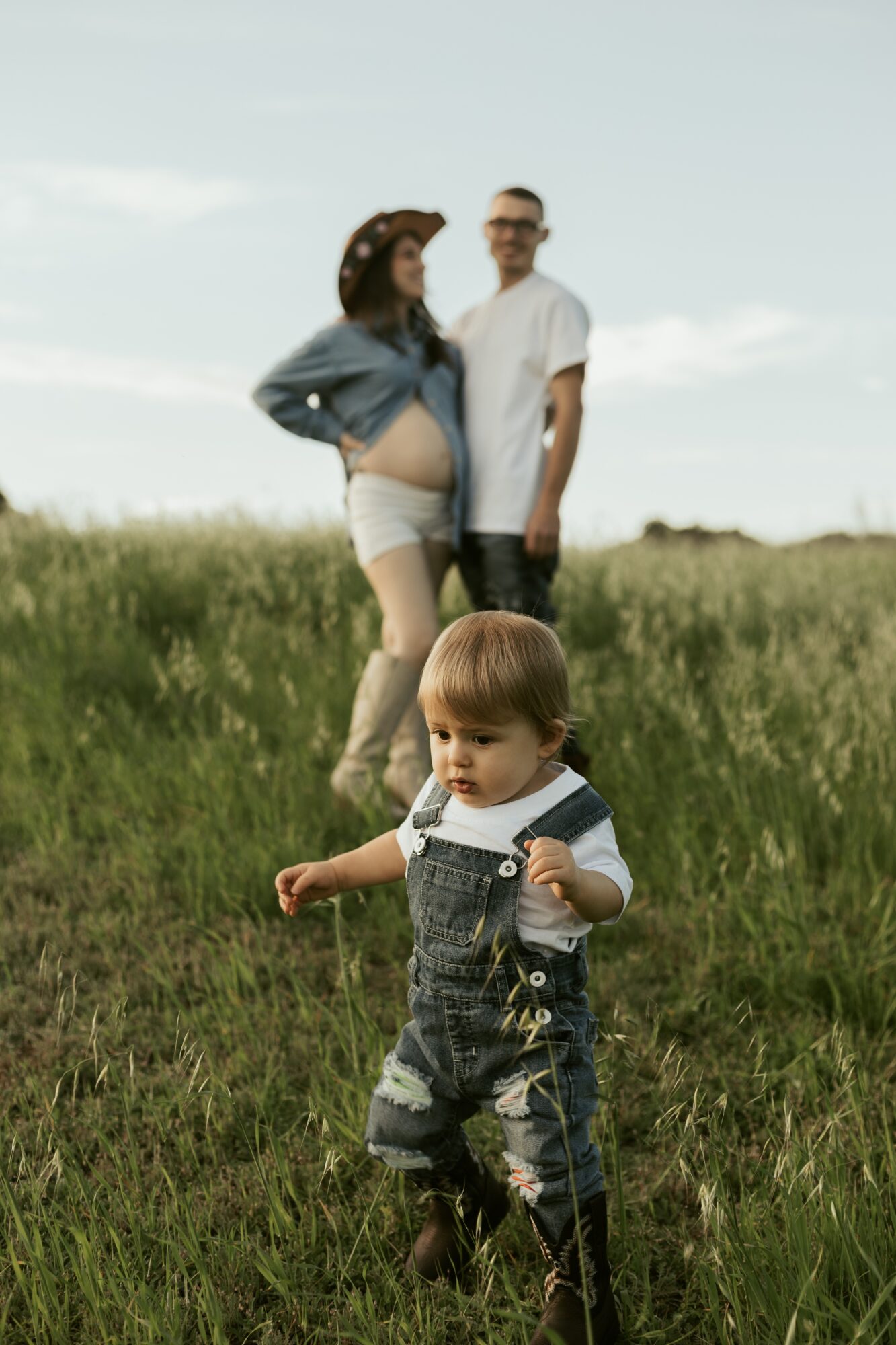 A young child walking in a grassy field with a couple in the background, one pregnant woman and a man, under a cloudy sky.