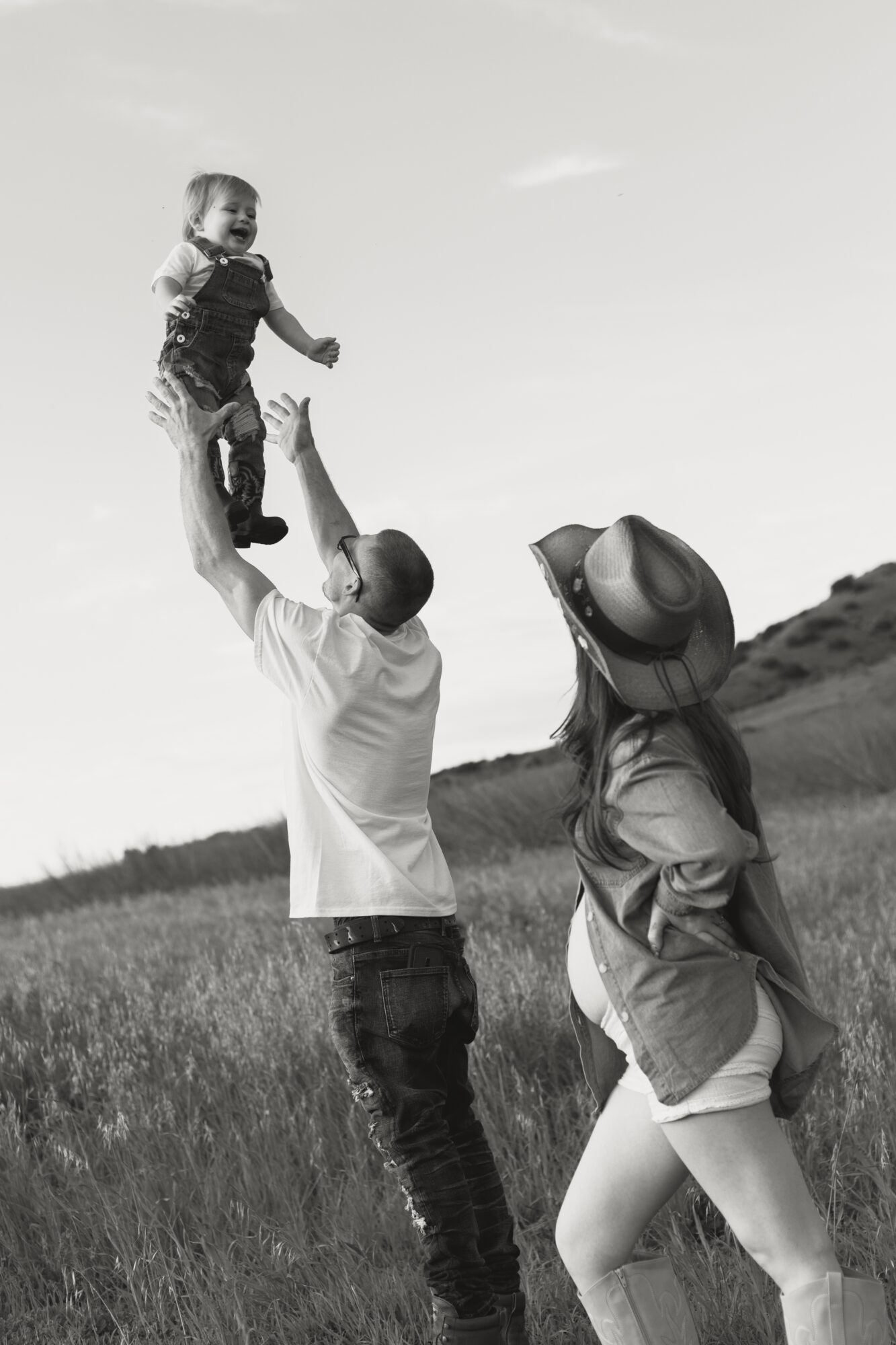 A man lifts a child into the air while a woman stands nearby in a grassy field, wearing a hat and looking up.
