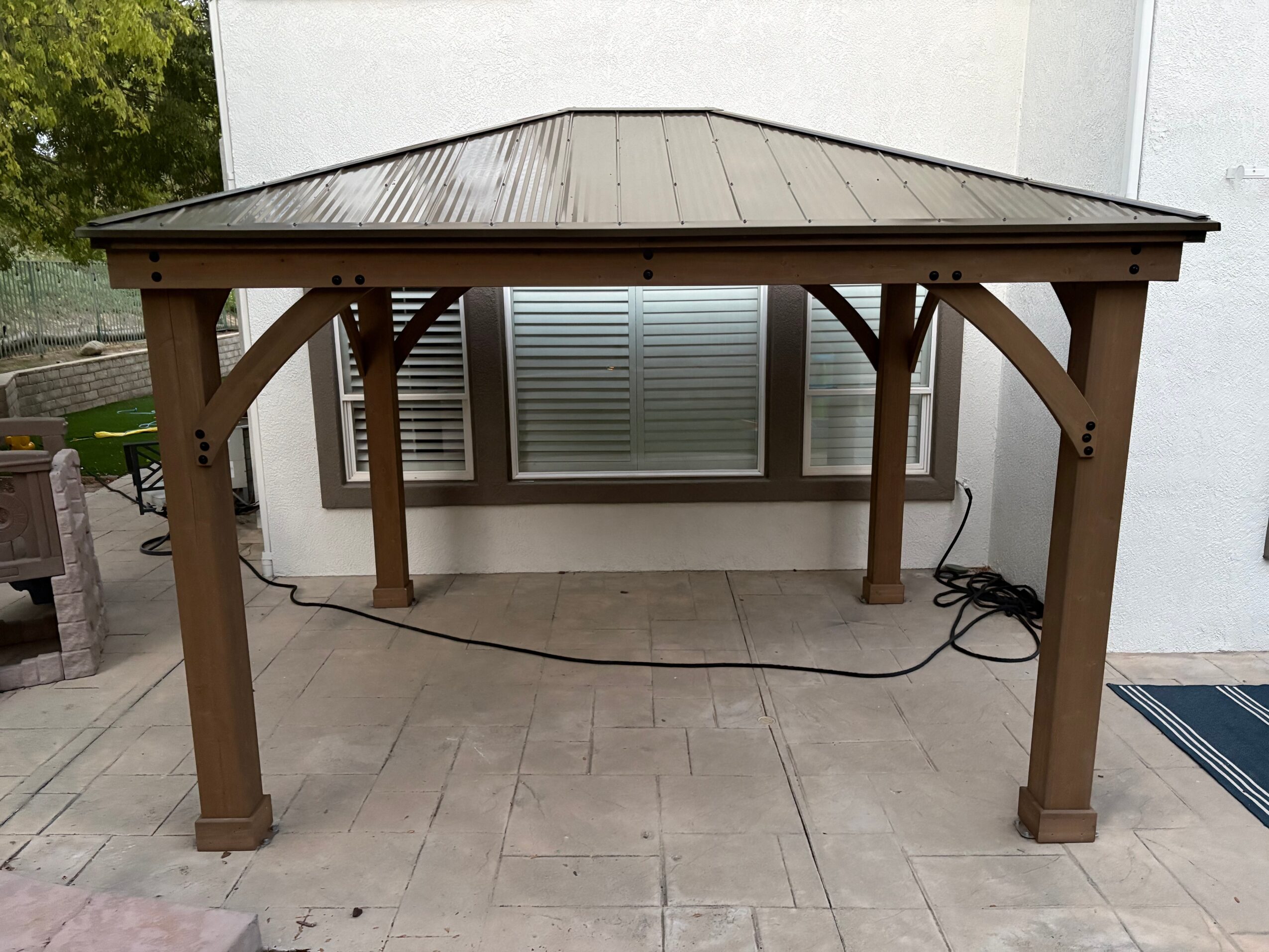 Wooden gazebo with metal roof on tiled patio, with window and wall in background.