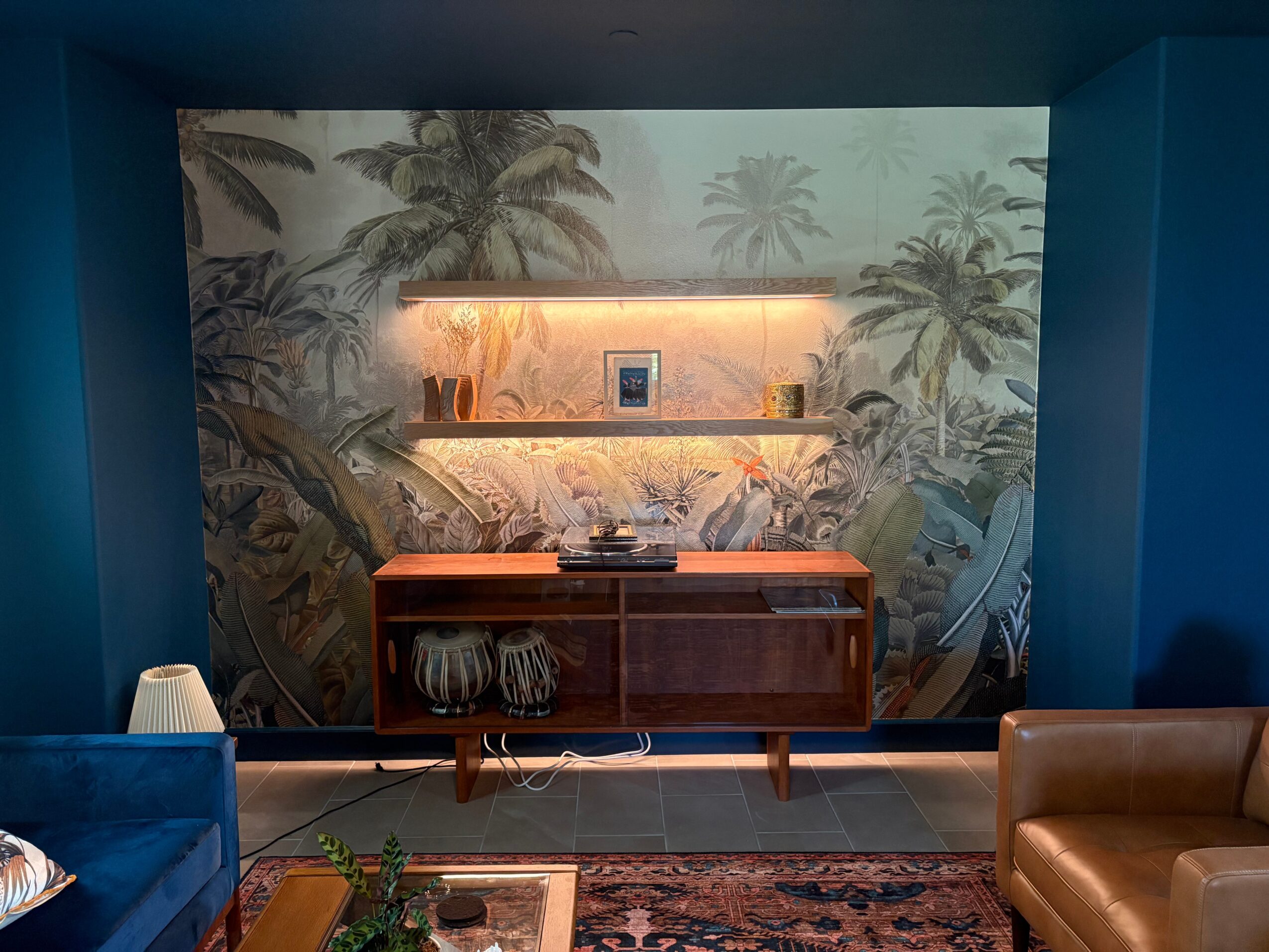 Decorative wall with tropical palm trees, a wooden sideboard, and a shelf with a small framed picture, illuminated by warm lighting.