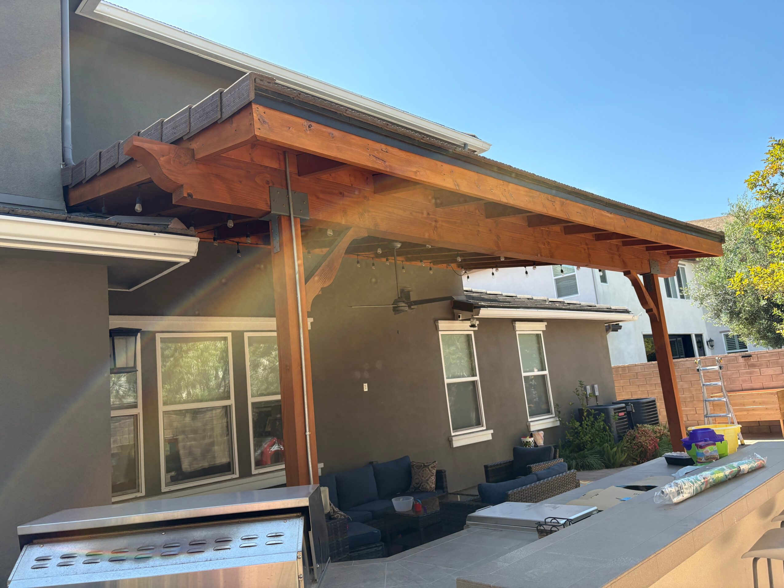Outdoor patio area with seating, covered by a wooden roof extension, adjacent to a gray house with windows and sliding door.