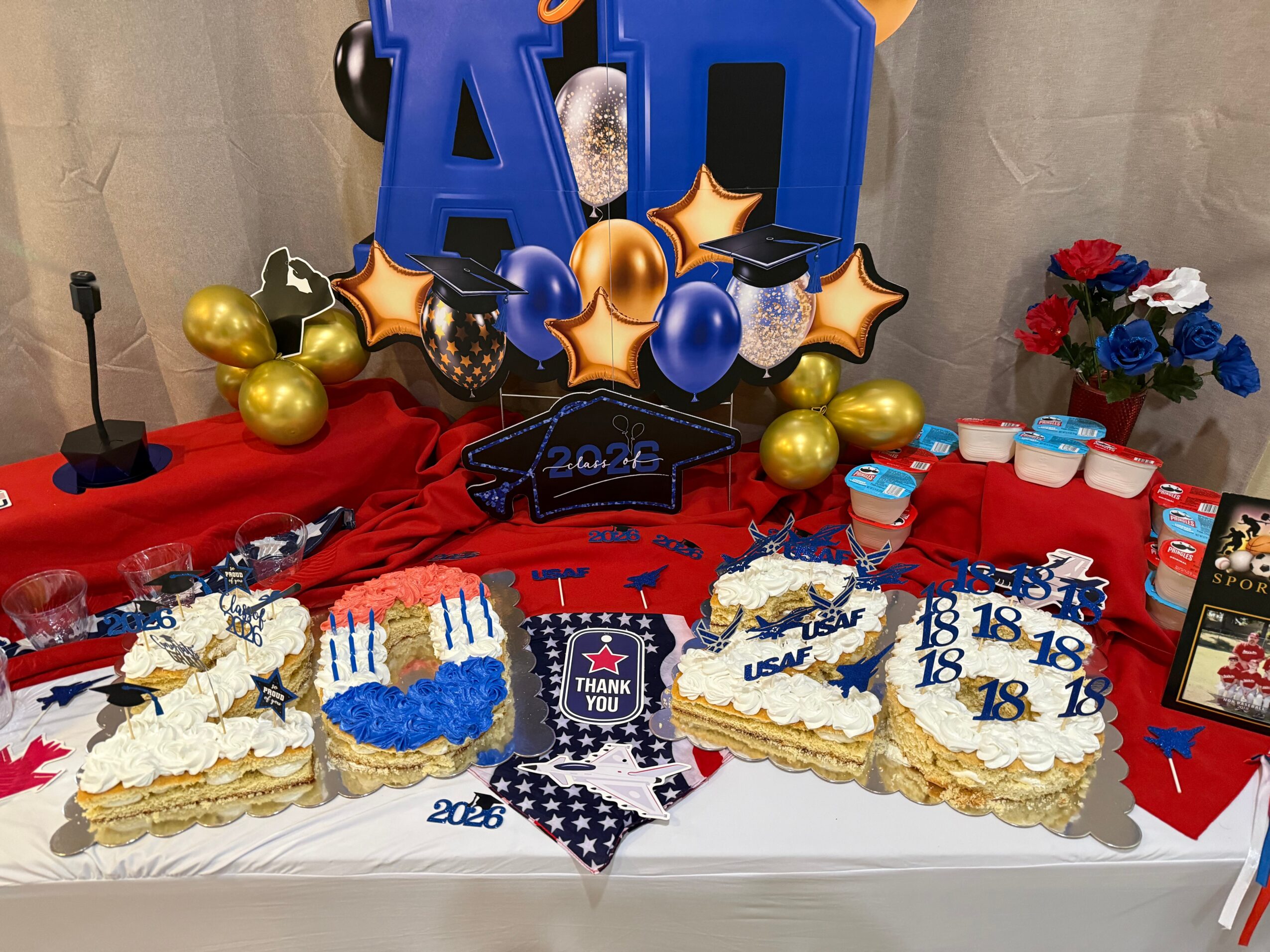 Celebration table with decorated cakes, balloons, and a bouquet of flowers, featuring a large blue and gold 'AID' sign and 2024 theme.