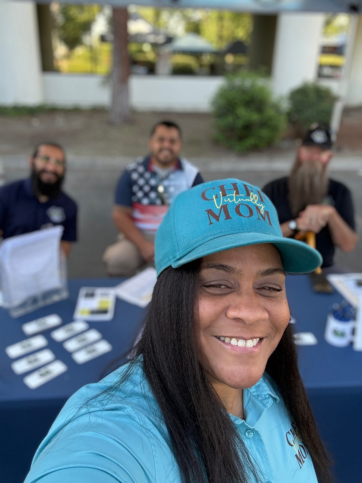 Woman smiling in foreground wearing a blue cap and shirt, three people sitting at a table in background outdoors.