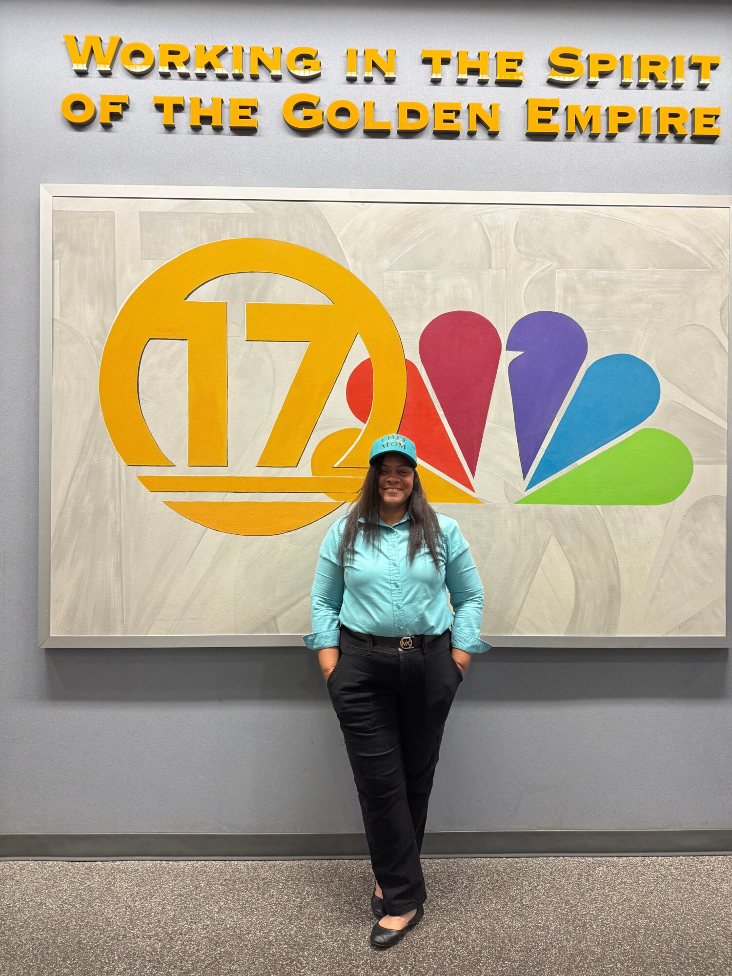 Woman in blue shirt and black pants standing in front of a colorful logo and text on a wall.