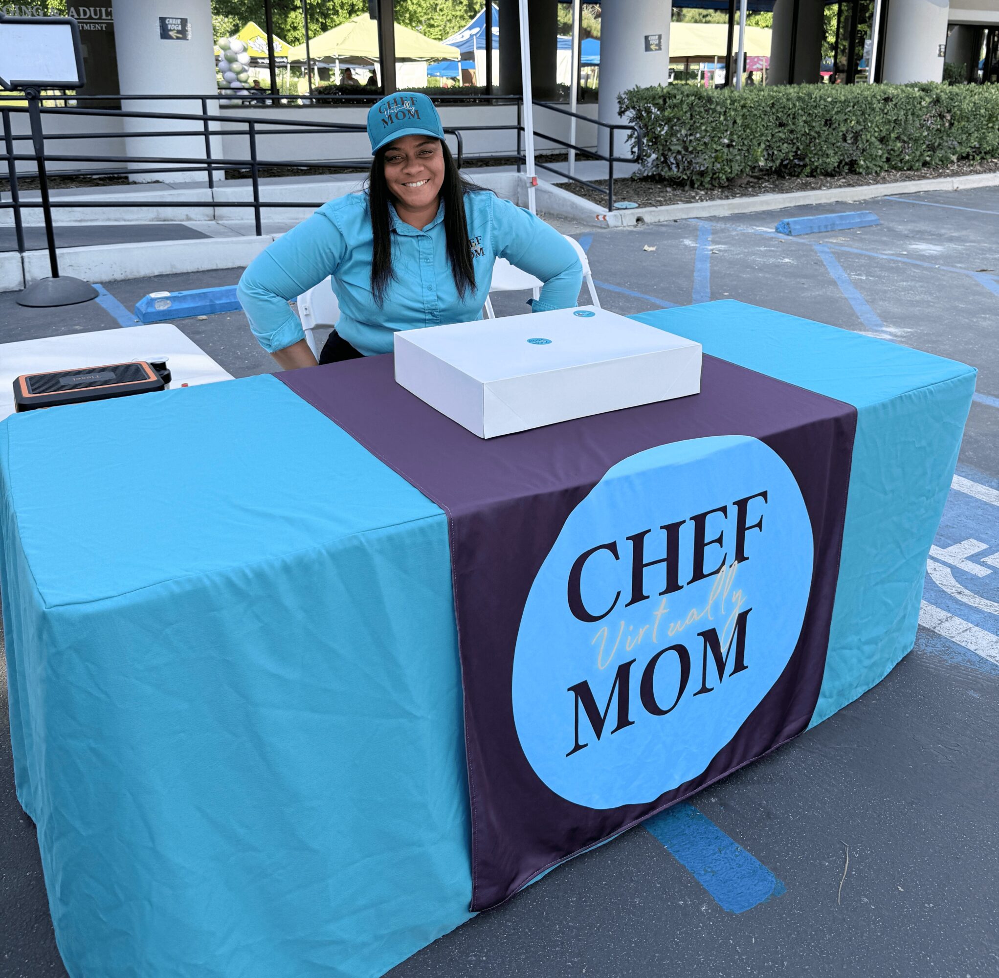 Smiling woman in blue shirt and cap sitting behind a table with a sign that says 'Chef Mom' outdoors.
