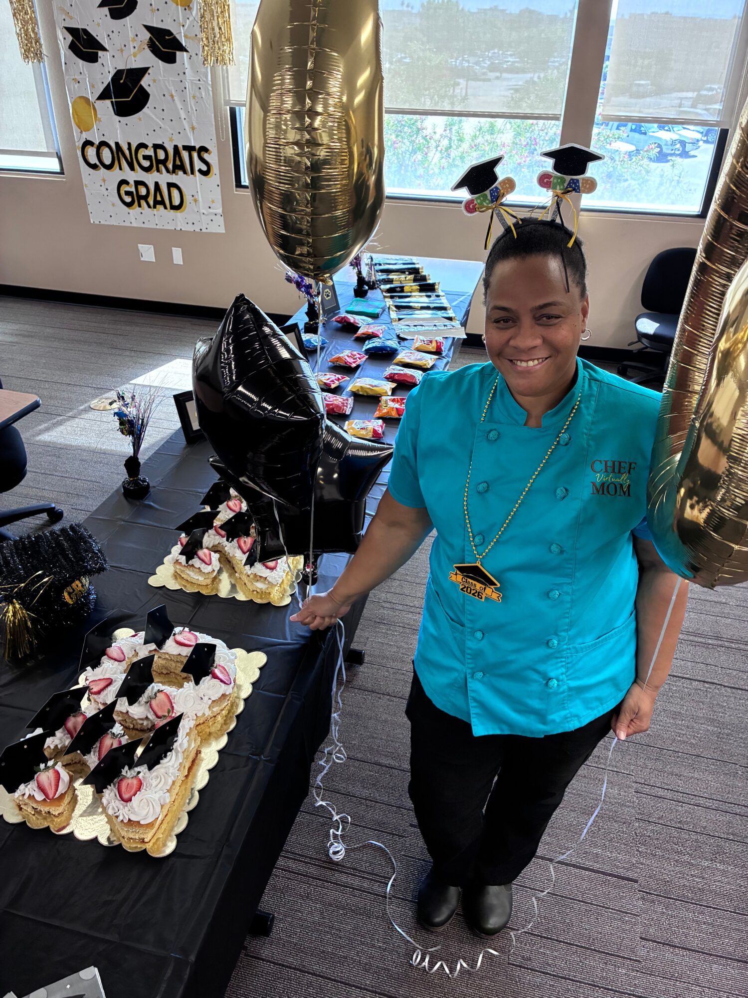 Woman in blue uniform standing next to a table with a decorated cake, balloons, and a congratulatory sign, celebrating an achievement.
