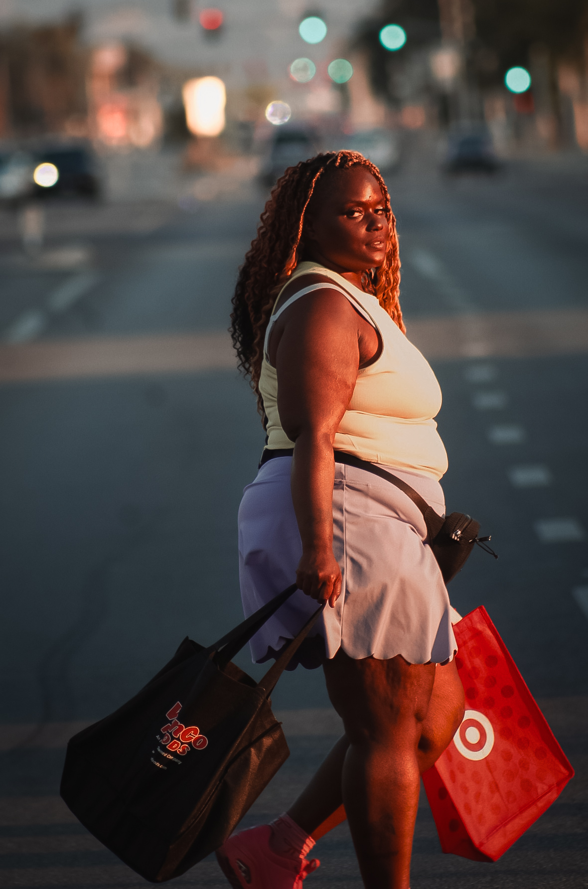 Woman with curly hair in a sleeveless top and skirt walking on street carrying shopping bags.