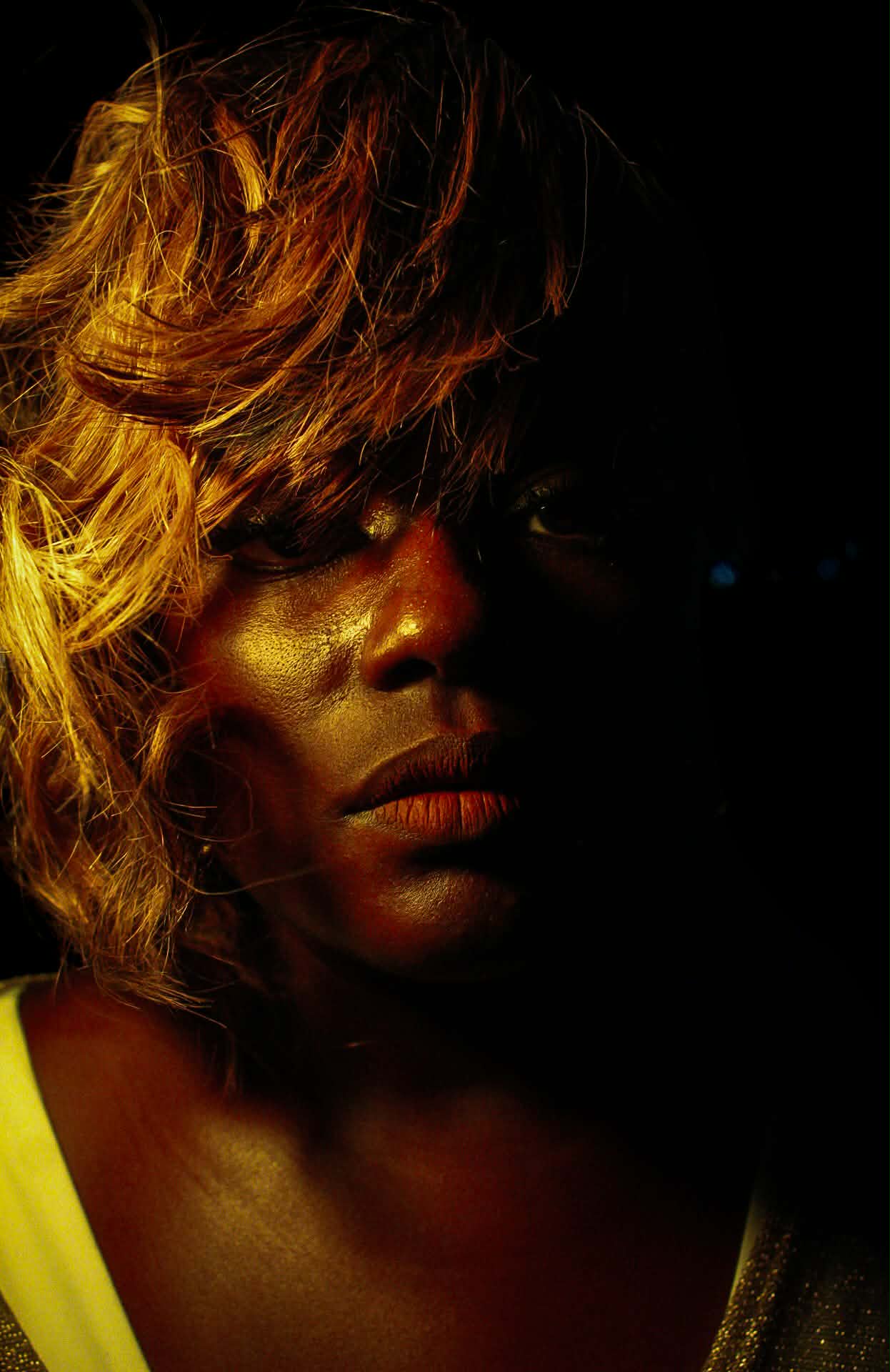 Portrait of a person with curly hair, partially illuminated, dark background, serious expression.