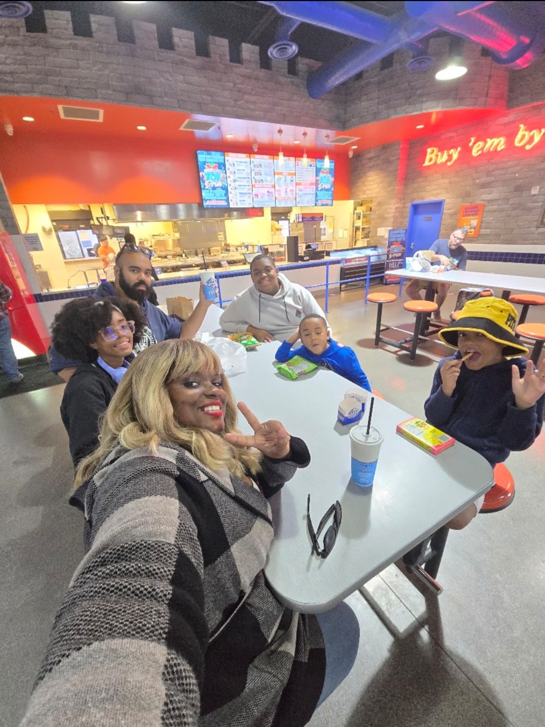 Group of six people sitting at a table inside a fast-food restaurant, smiling and posing for a selfie, with menu boards and counter in background.