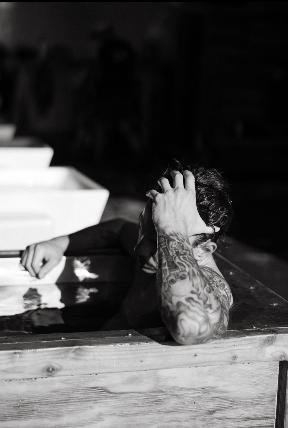 Person with tattoos resting head on arm at a table, hand in hair, in black and white.