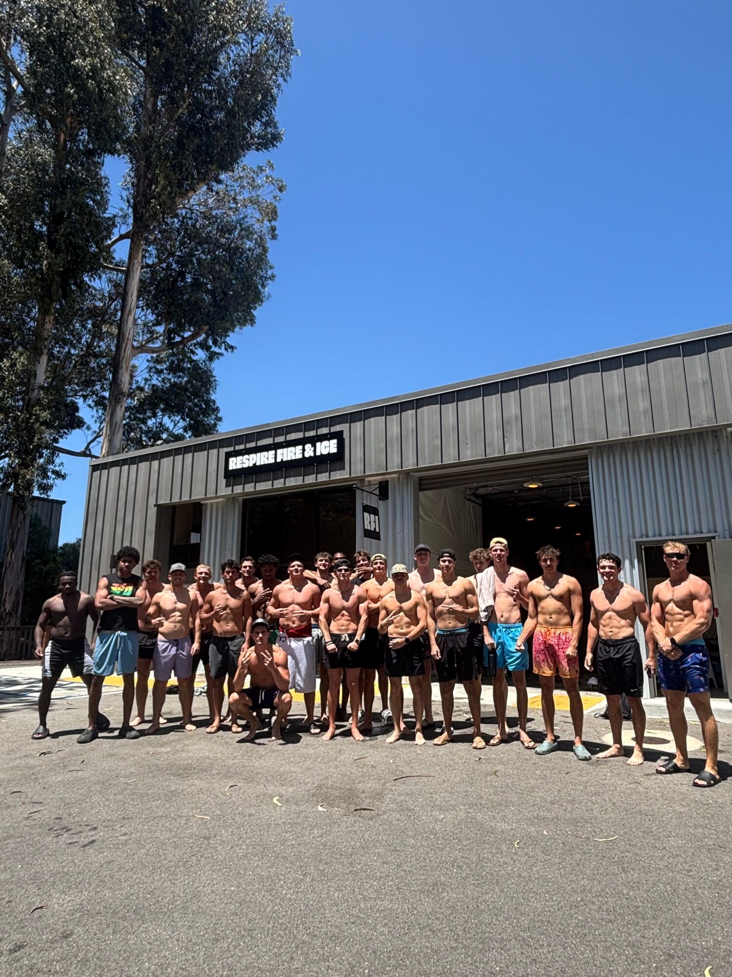 Group of people standing outside a building, some shirtless, with trees and a clear blue sky in the background.