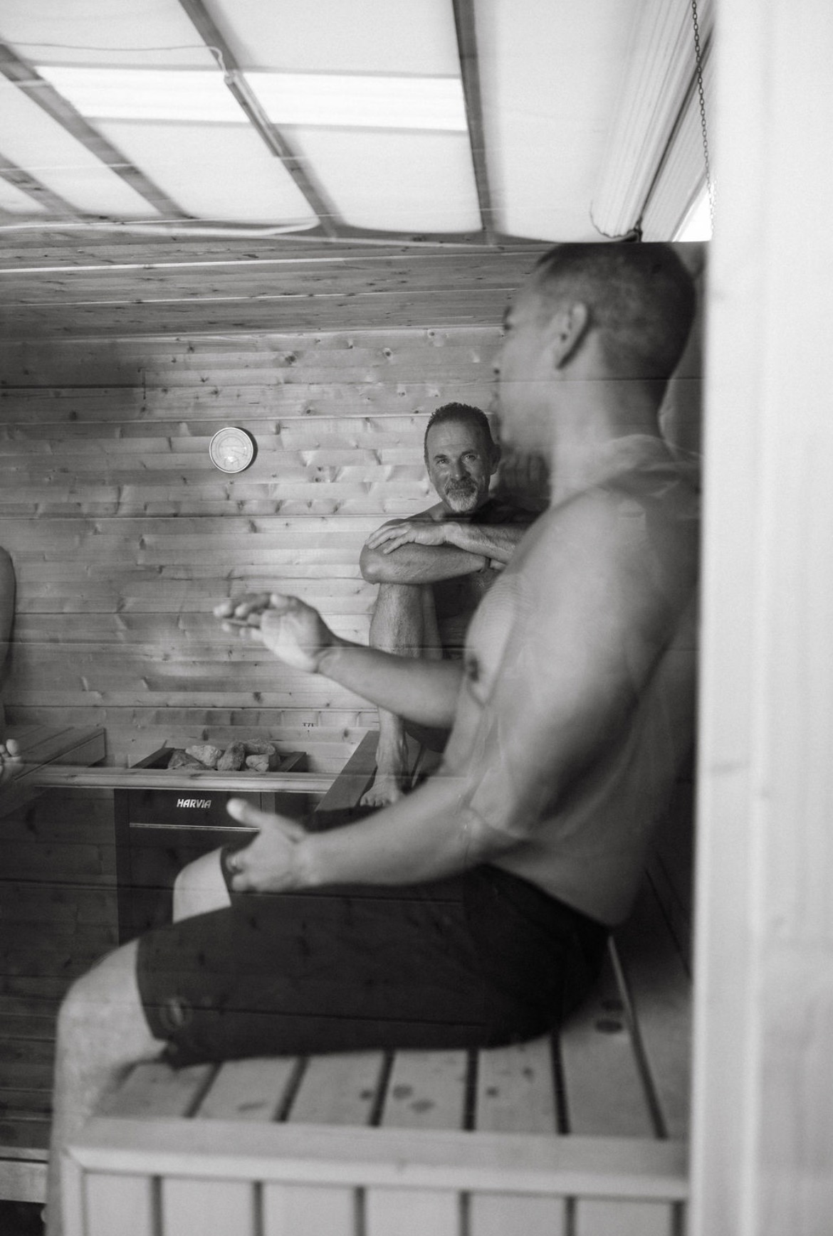 A shirtless man sitting on a bench inside a wooden sauna, with another person standing behind him, smiling.