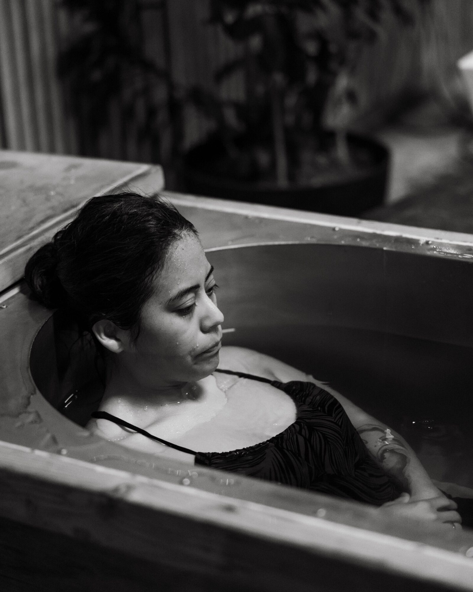 Woman with dark hair in a bun relaxing in a wooden tub, eyes closed, in a dimly lit room.