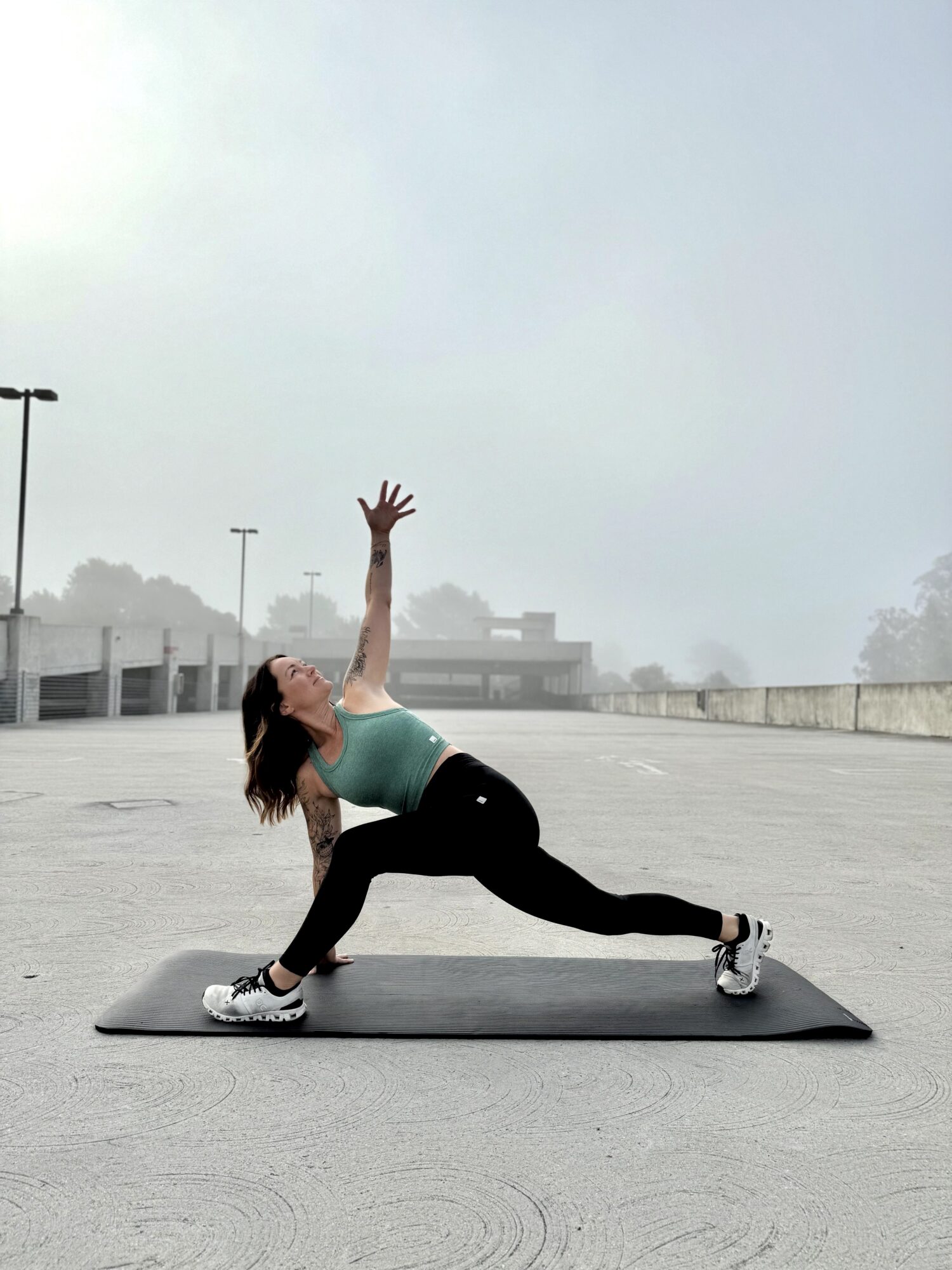Woman performing a yoga pose on a mat outdoors, with an overcast sky and parking lot in background.