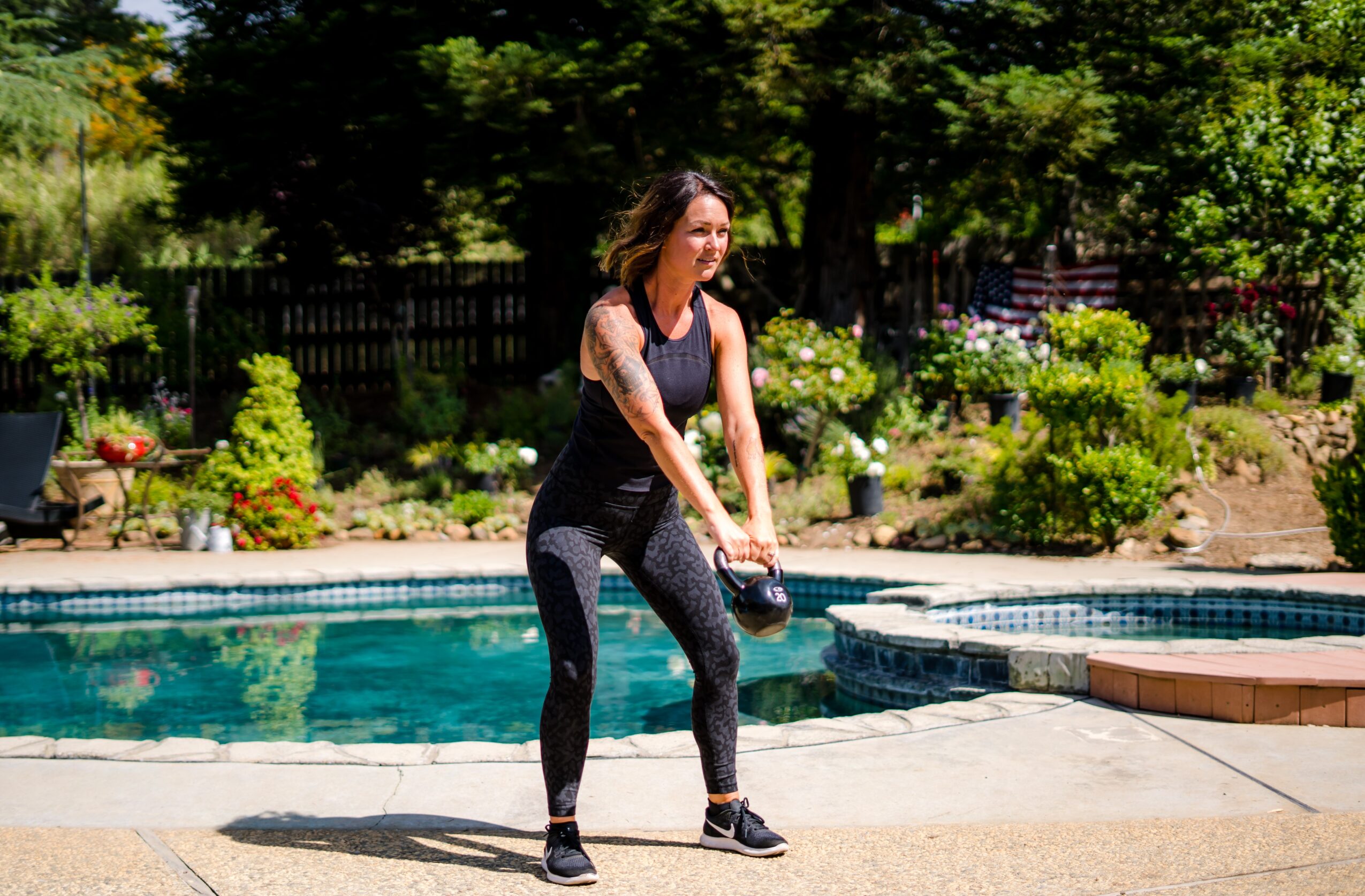 Woman in black outfit holding a ball near a swimming pool, surrounded by greenery and trees.
