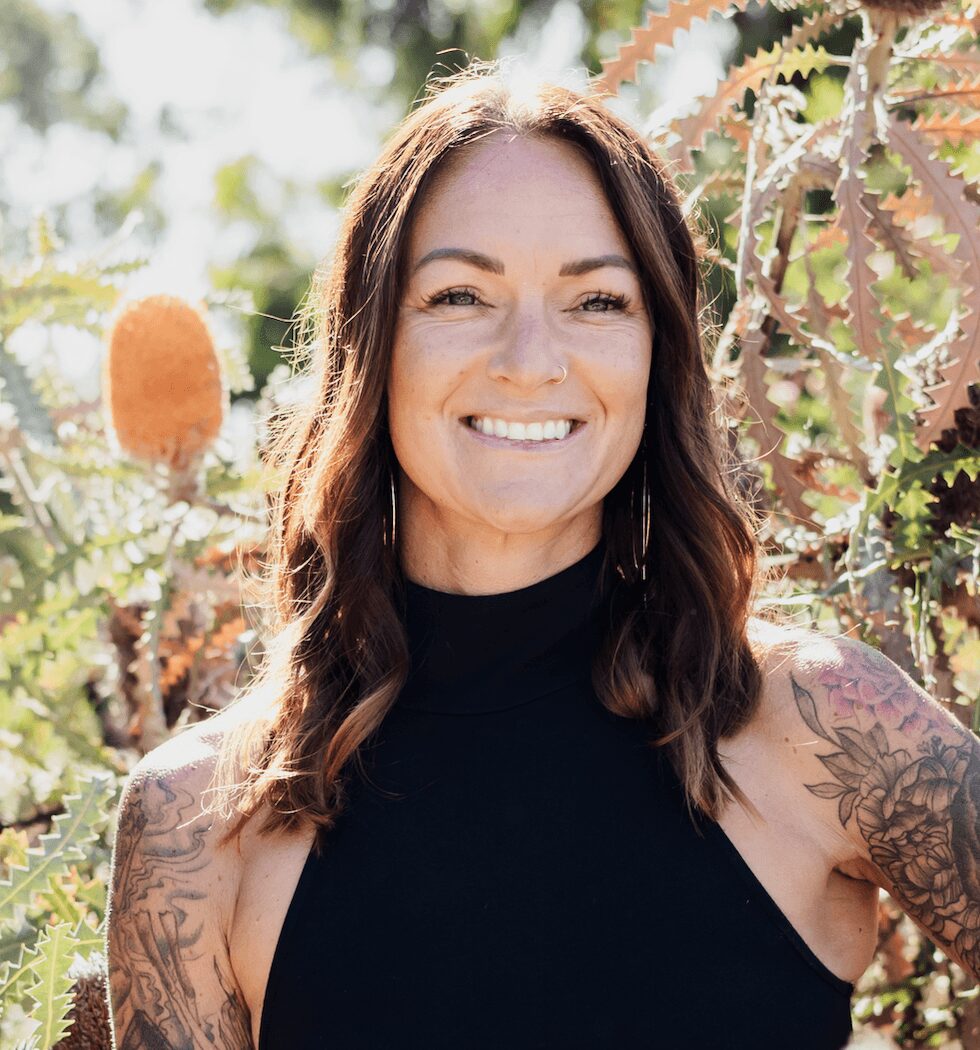 Woman with long brown hair smiling outdoors among plants, wearing a black sleeveless top.