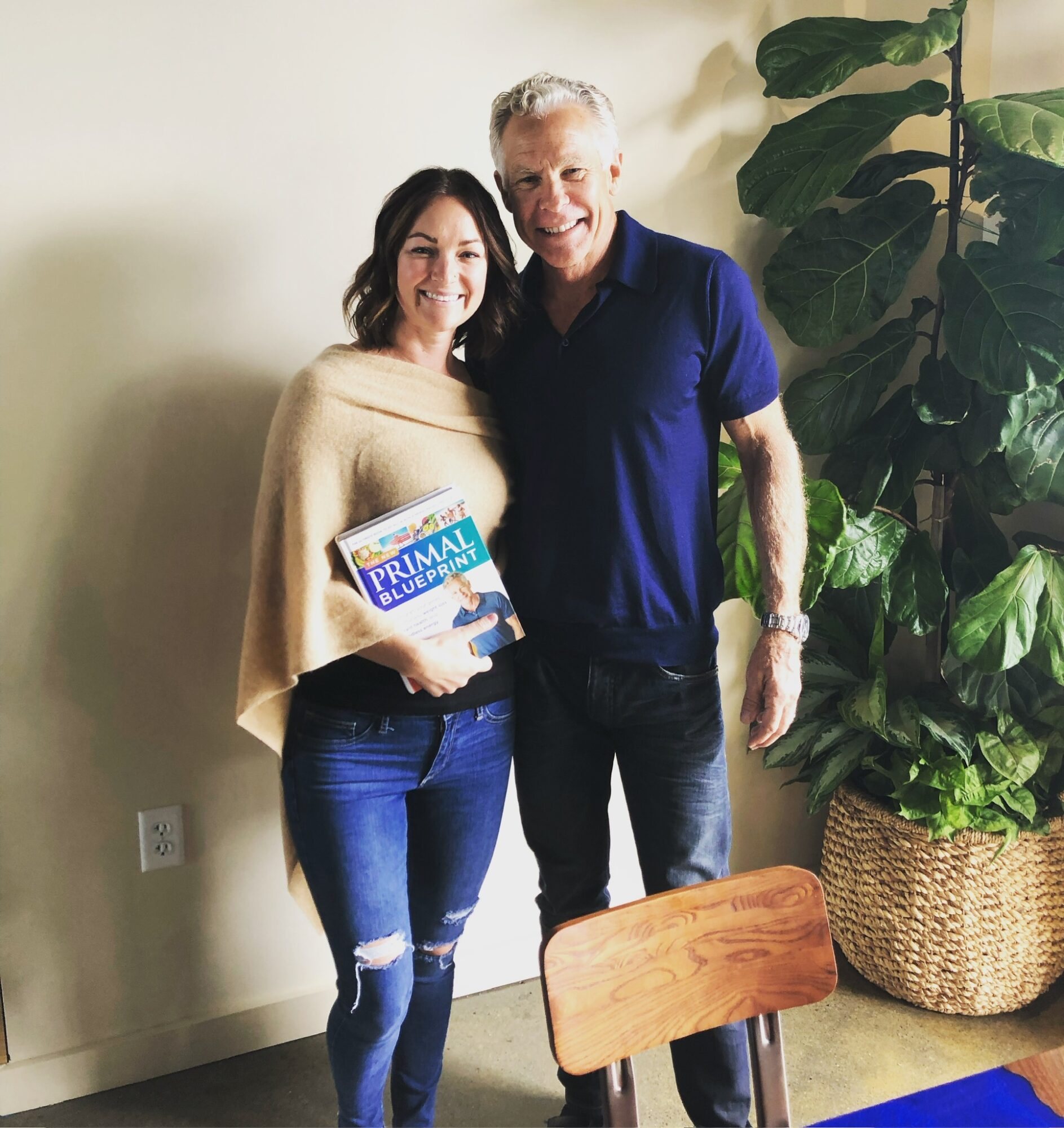 Two people standing indoors, smiling, with a large plant in a basket nearby, one holding a book titled 'PRIMAL BLEND'.