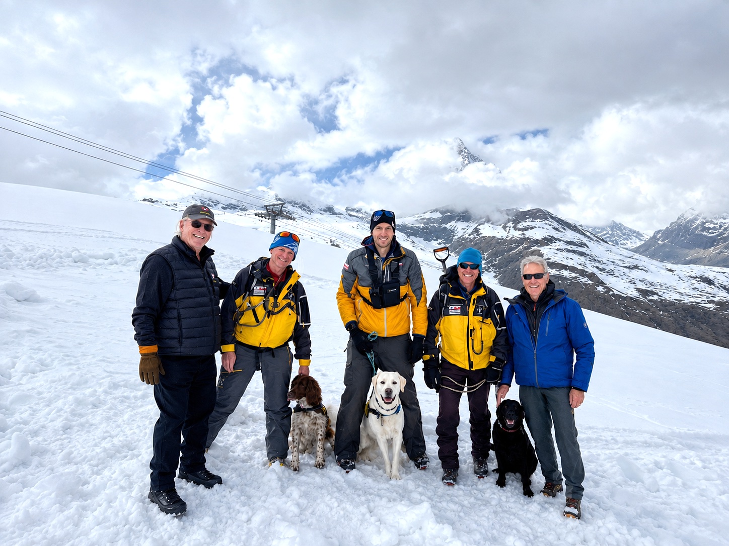 Group of five people with three dogs standing on snowy mountain landscape with cloudy sky and mountain peaks in background.