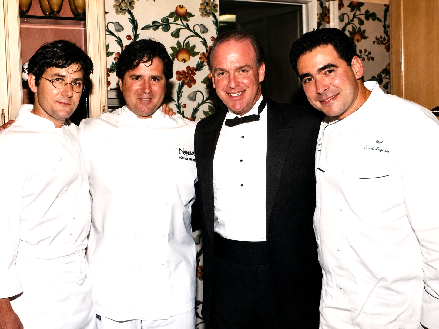 Four men, two in chef uniforms and two in formal attire, standing together indoors with floral wallpaper background.