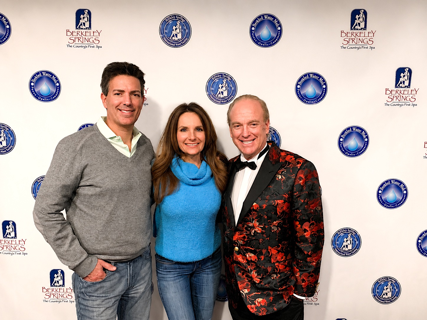Three people standing together in front of a backdrop with logos, smiling, with two men and one woman, all dressed casually and formally.