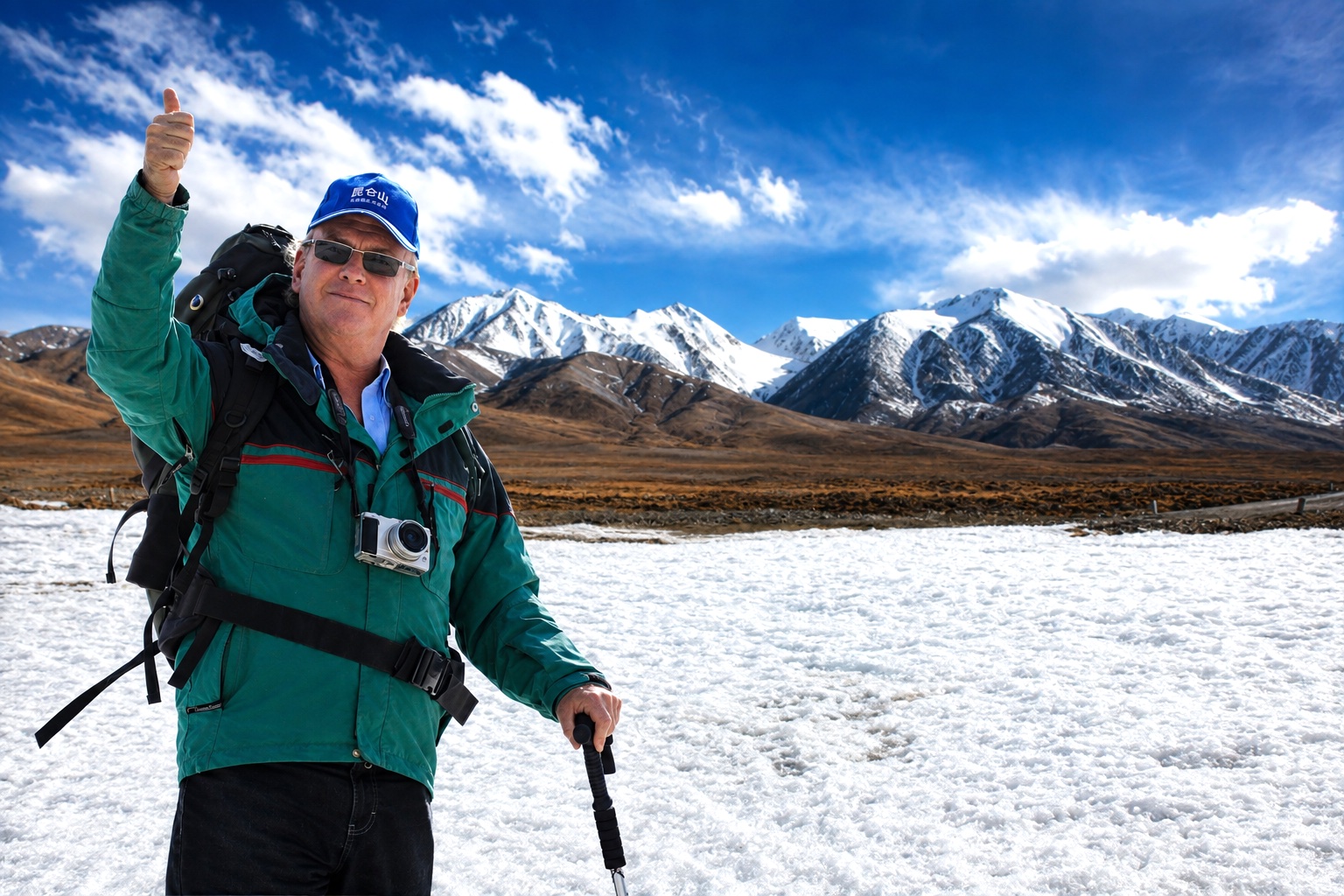 Man in outdoor gear holding a walking stick with snowy mountains and blue sky in background.