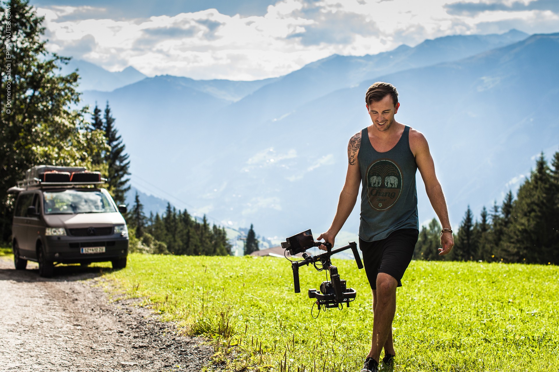 Person walking on grassy field with mountains and trees in background, holding a drone controller, sunny day.