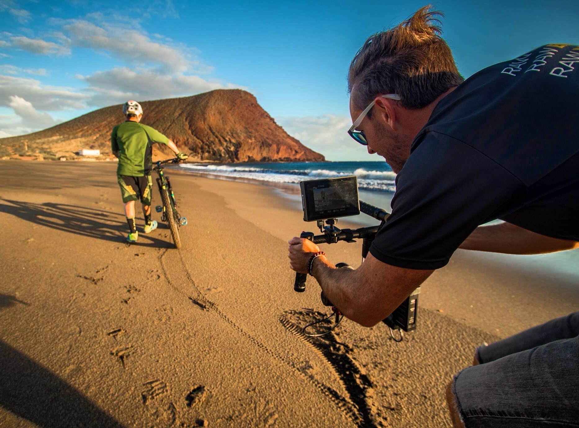Person filming another person walking on a beach with a mountain in the background, during sunset or sunrise.