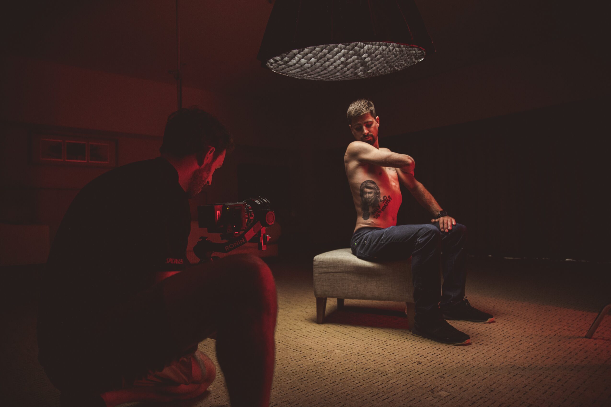 Person sitting on a bench with arms crossed, tattooed, in a dimly lit room, with a photographer taking their picture