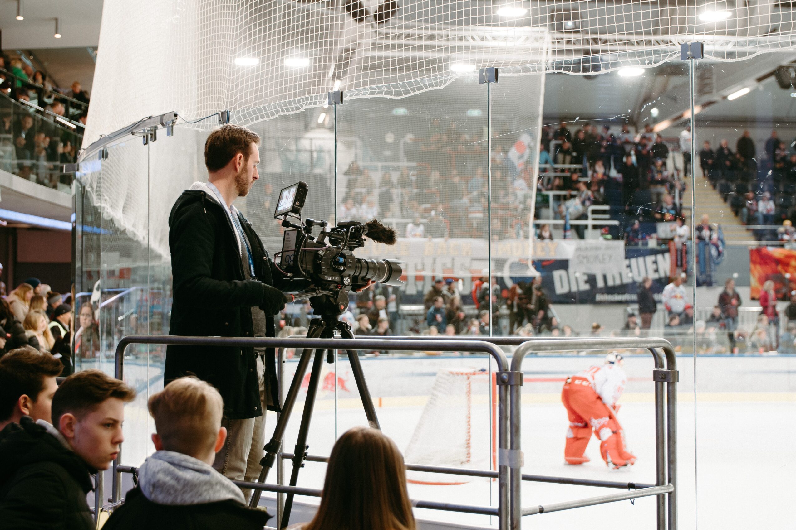 A cameraman films a hockey game in an indoor arena with spectators, a player on ice, and a protective glass barrier.