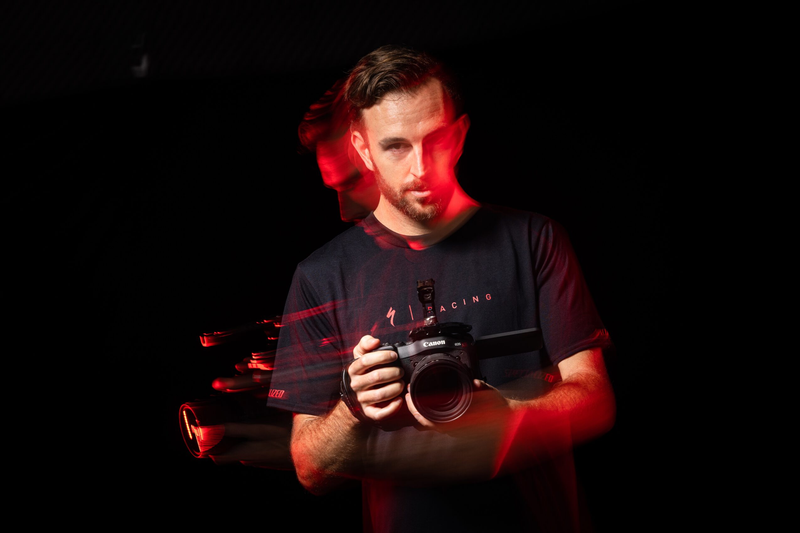 Man with beard holding camera, dark background, red lighting, looking at camera, casual clothing.