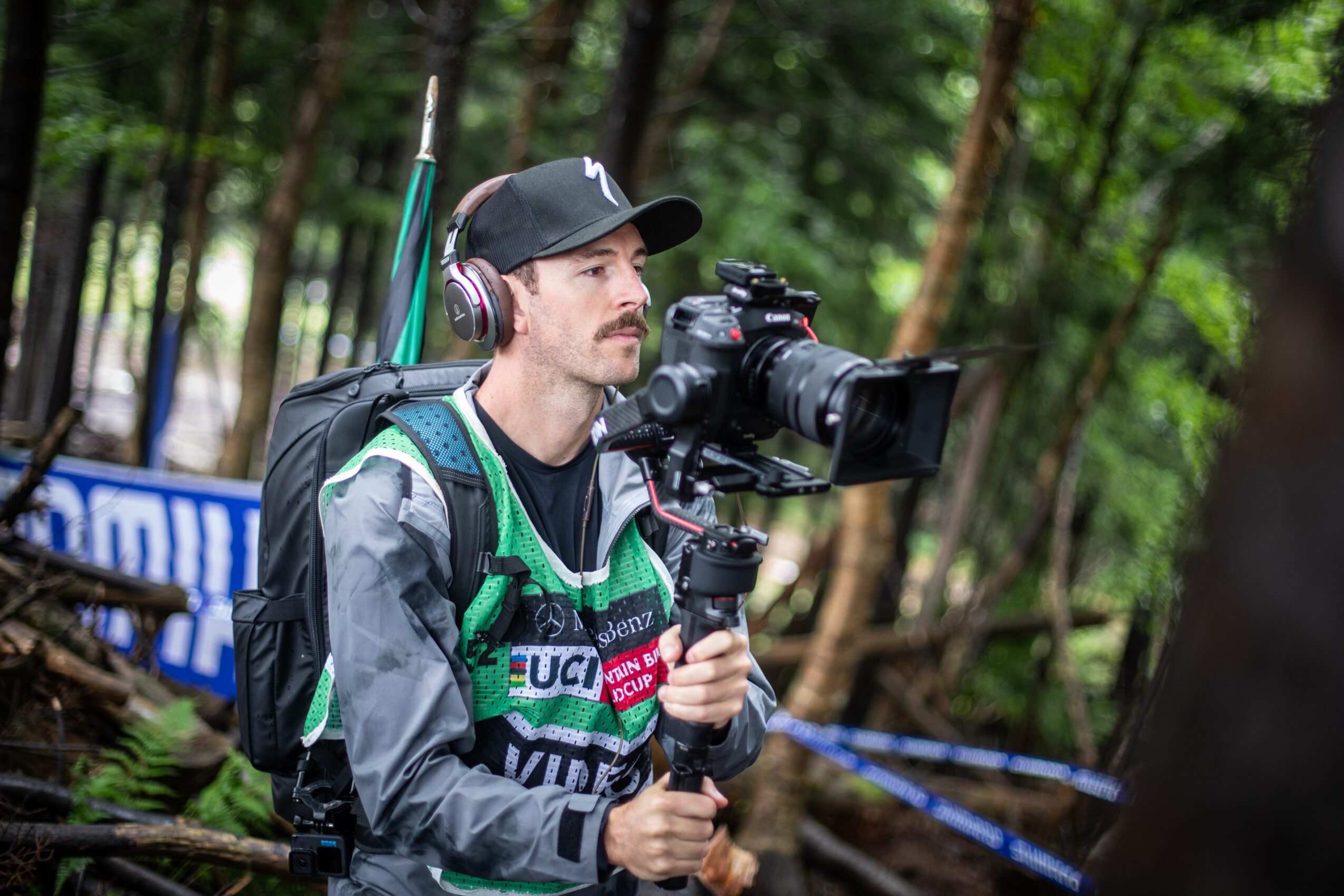 Man with headphones and a cap operating a camera on a stabilizer in a forest setting.