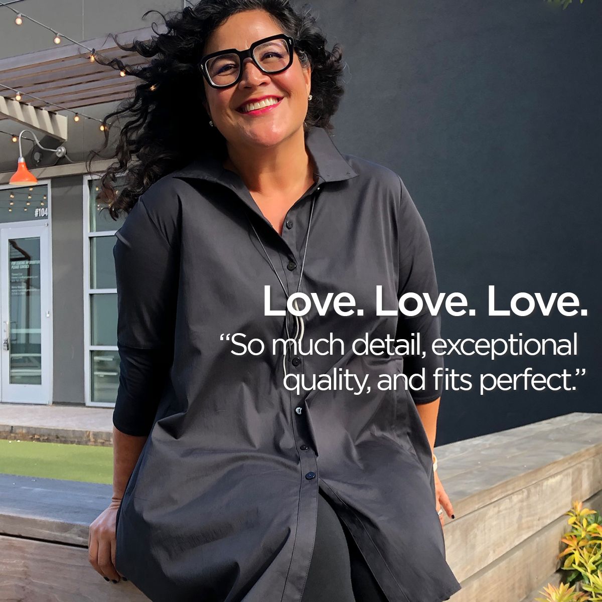 Smiling woman with glasses and curly hair wearing a dark dress outdoors, with a building and plants in background.