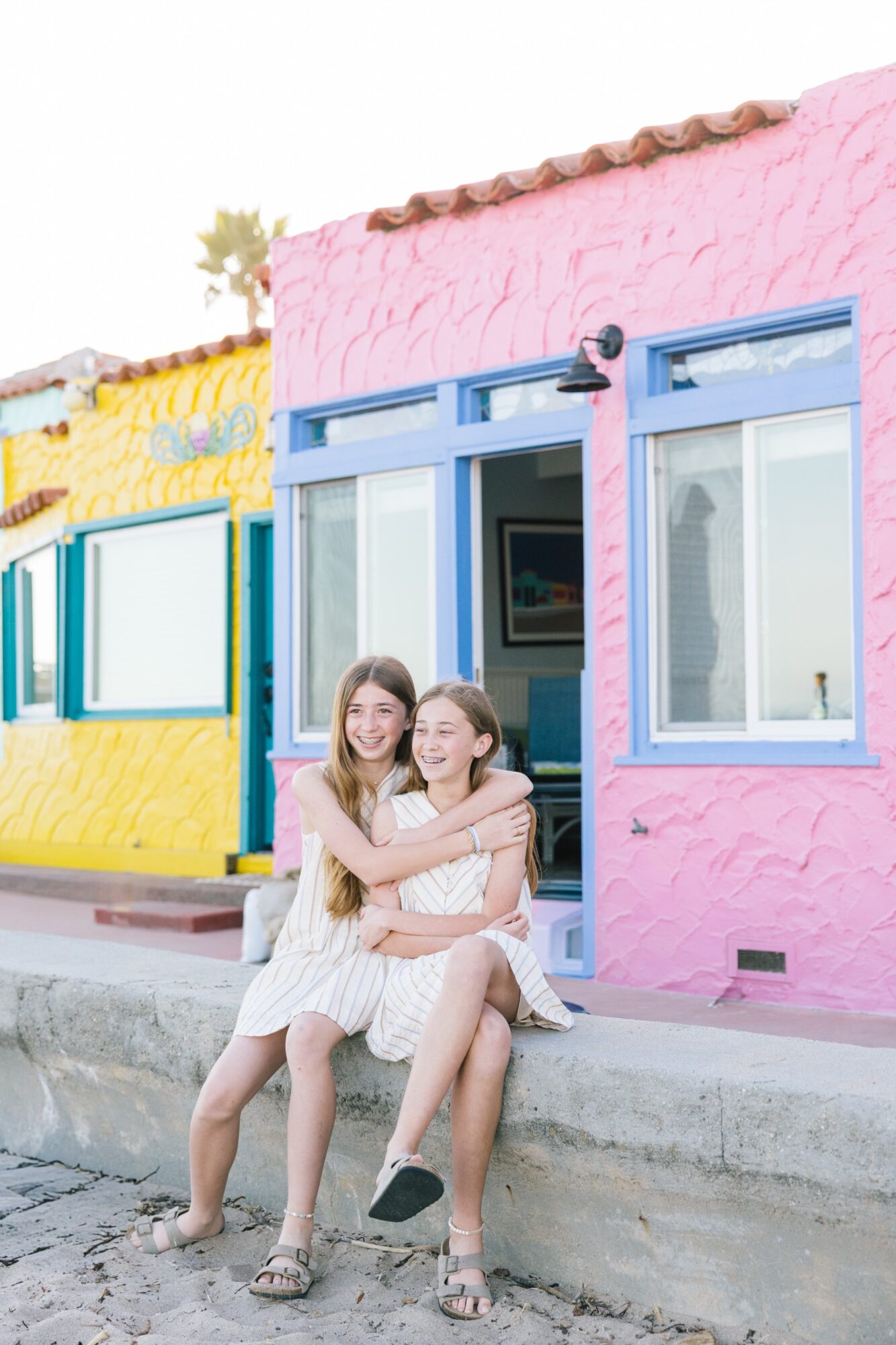 Two young women sit on a sidewalk in front of colorful buildings, smiling and hugging each other, with bright pink and yellow walls.