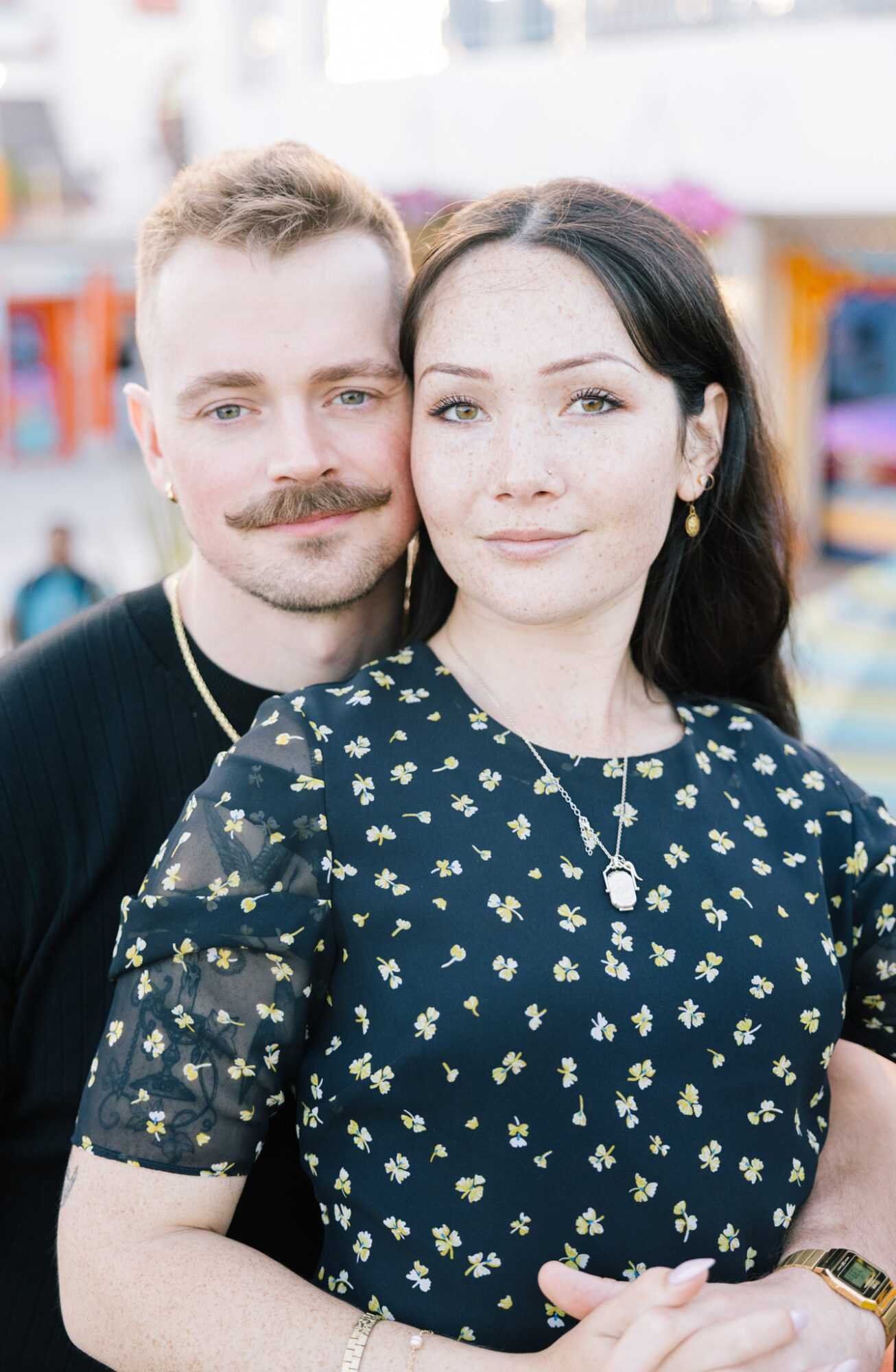 A man and woman pose closely outdoors, smiling, with blurred background of colorful objects and people.