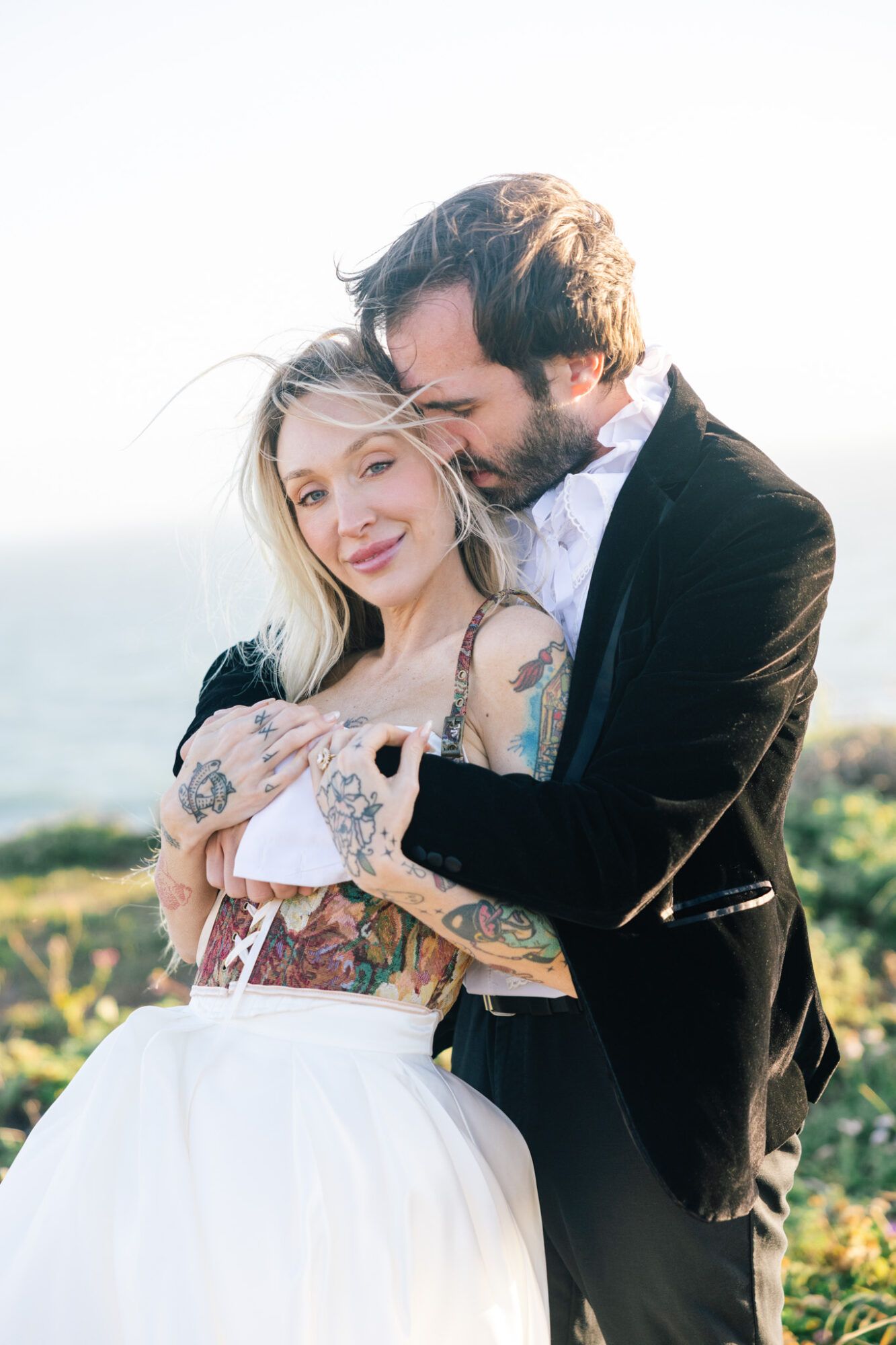 A man and woman embrace outdoors, smiling, with a scenic background. The woman has tattoos and blonde hair, the man has dark hair and a beard.