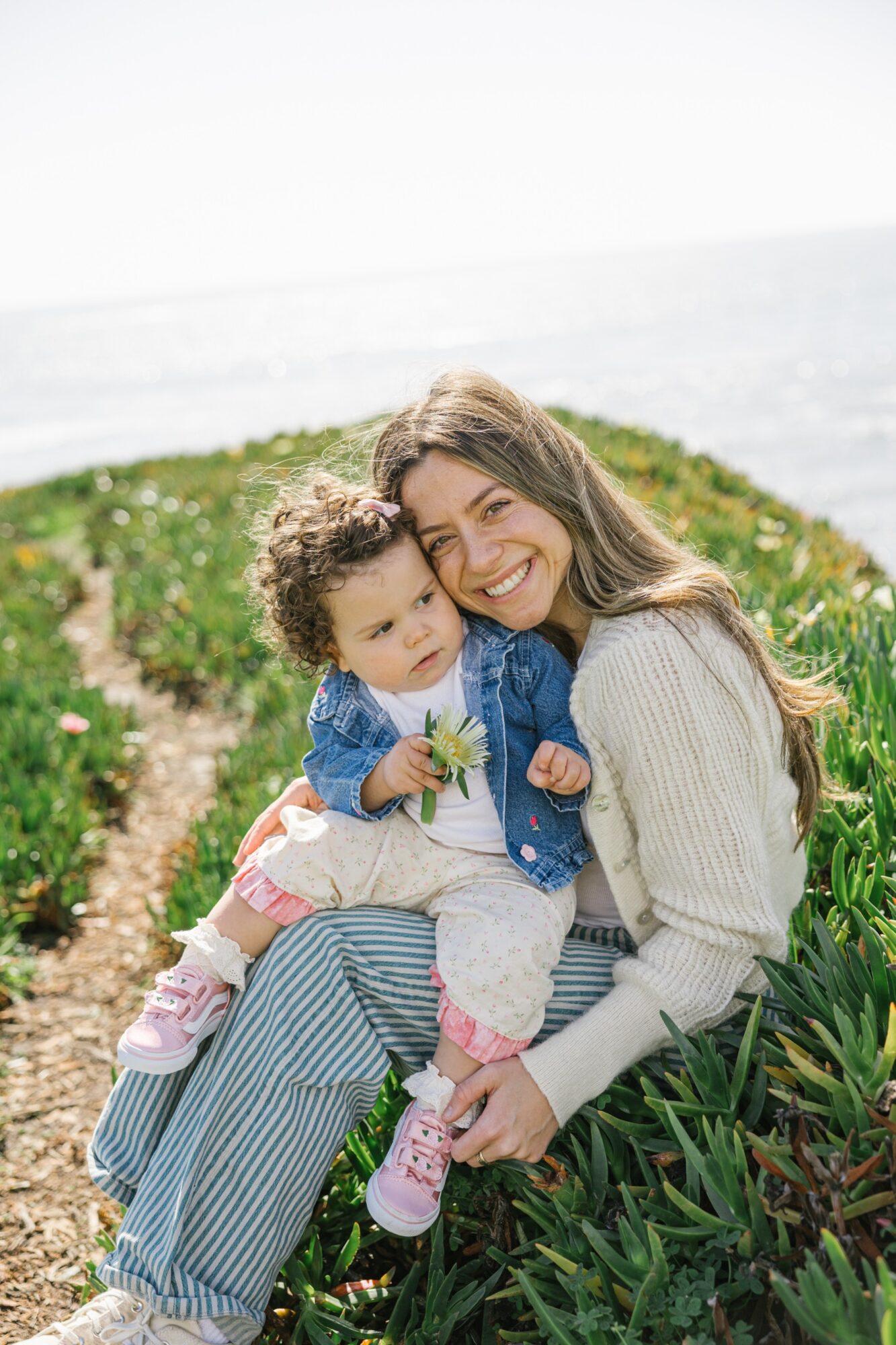 Woman holding a child outdoors near the ocean, both smiling, with greenery around them.