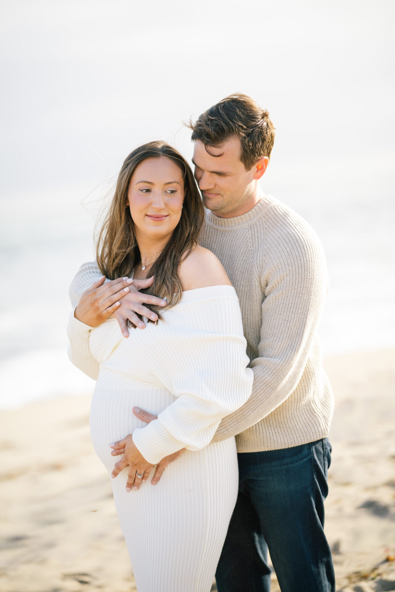 A couple stands on a beach, with the woman pregnant, embracing each other and smiling softly, bright sky in background.