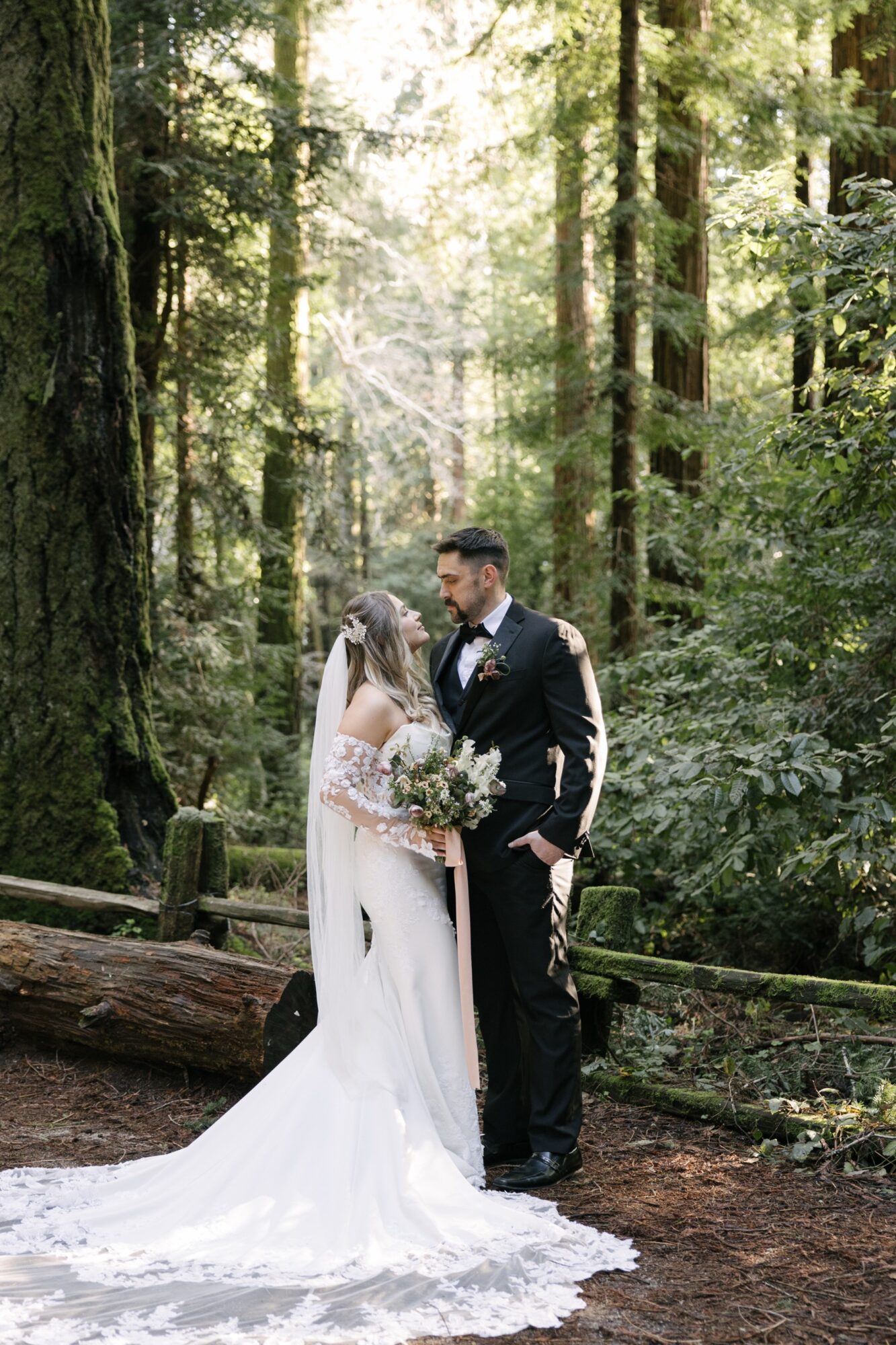 A bride and groom stand close together in a forest, holding a bouquet, surrounded by tall trees and greenery.