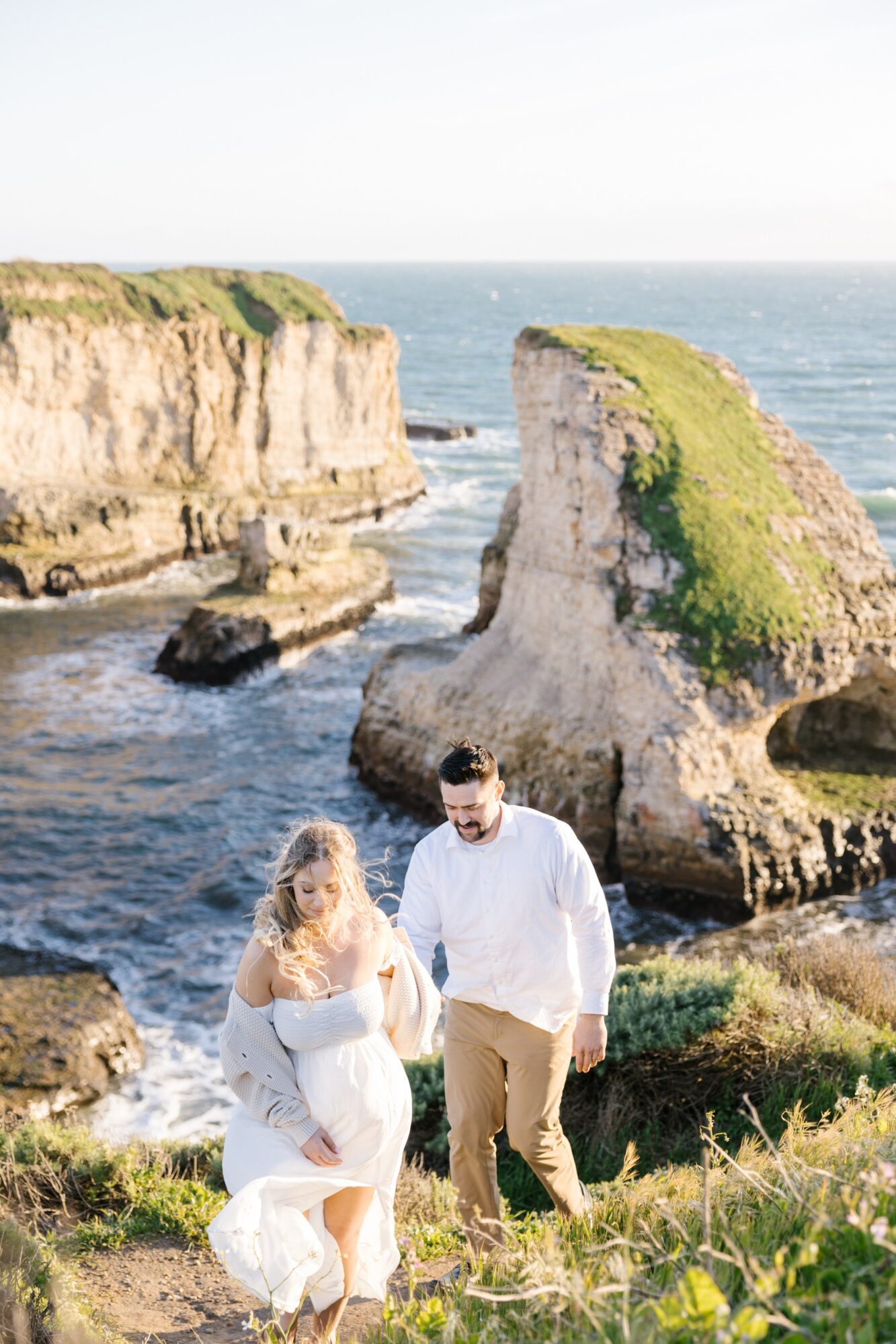 A couple walking along a rocky coastline with cliffs and ocean in the background, grassy area in foreground, sunny day.