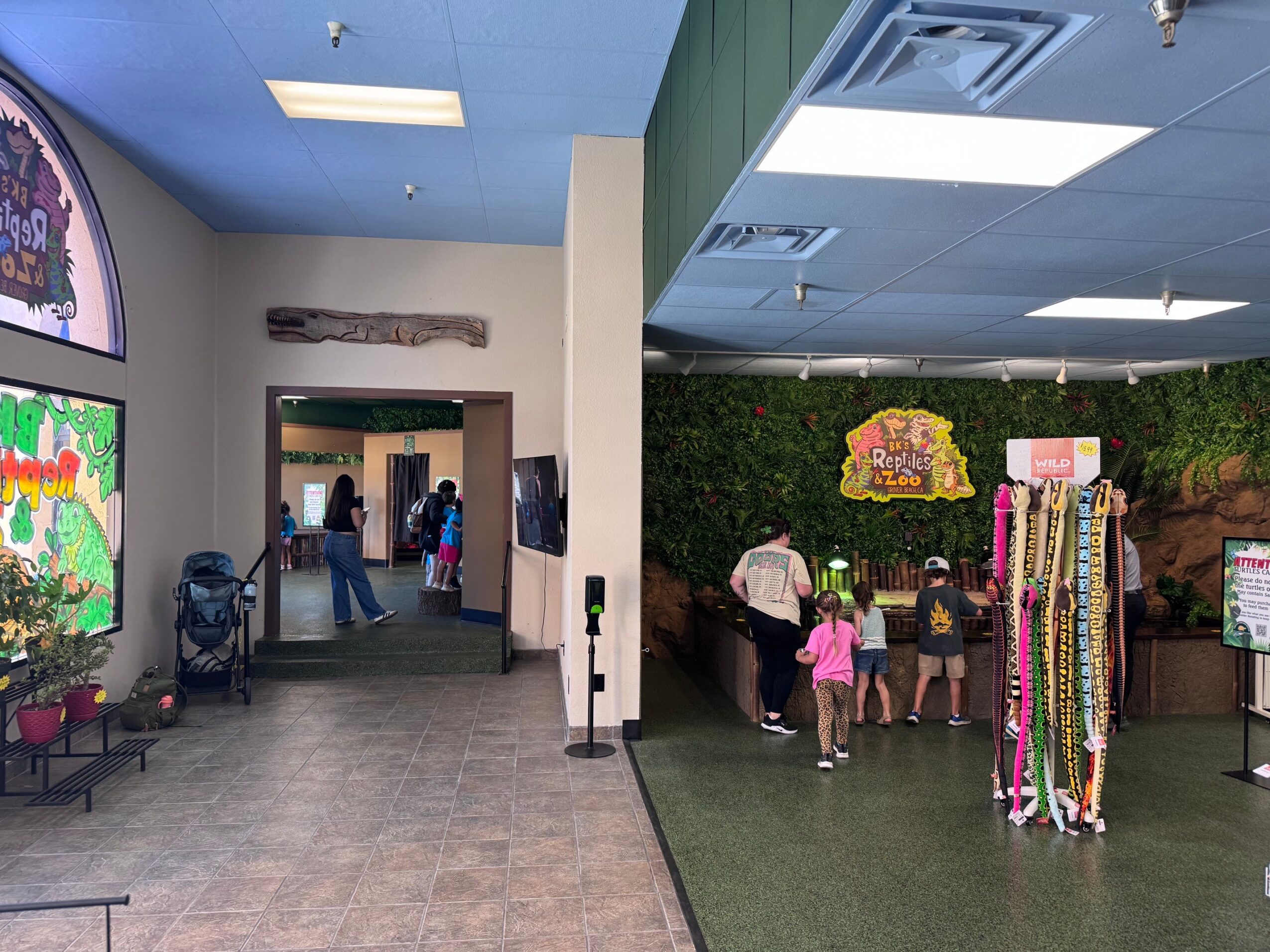 Indoor area with children and adults near a colorful display and a green wall, with a hallway leading to another room.