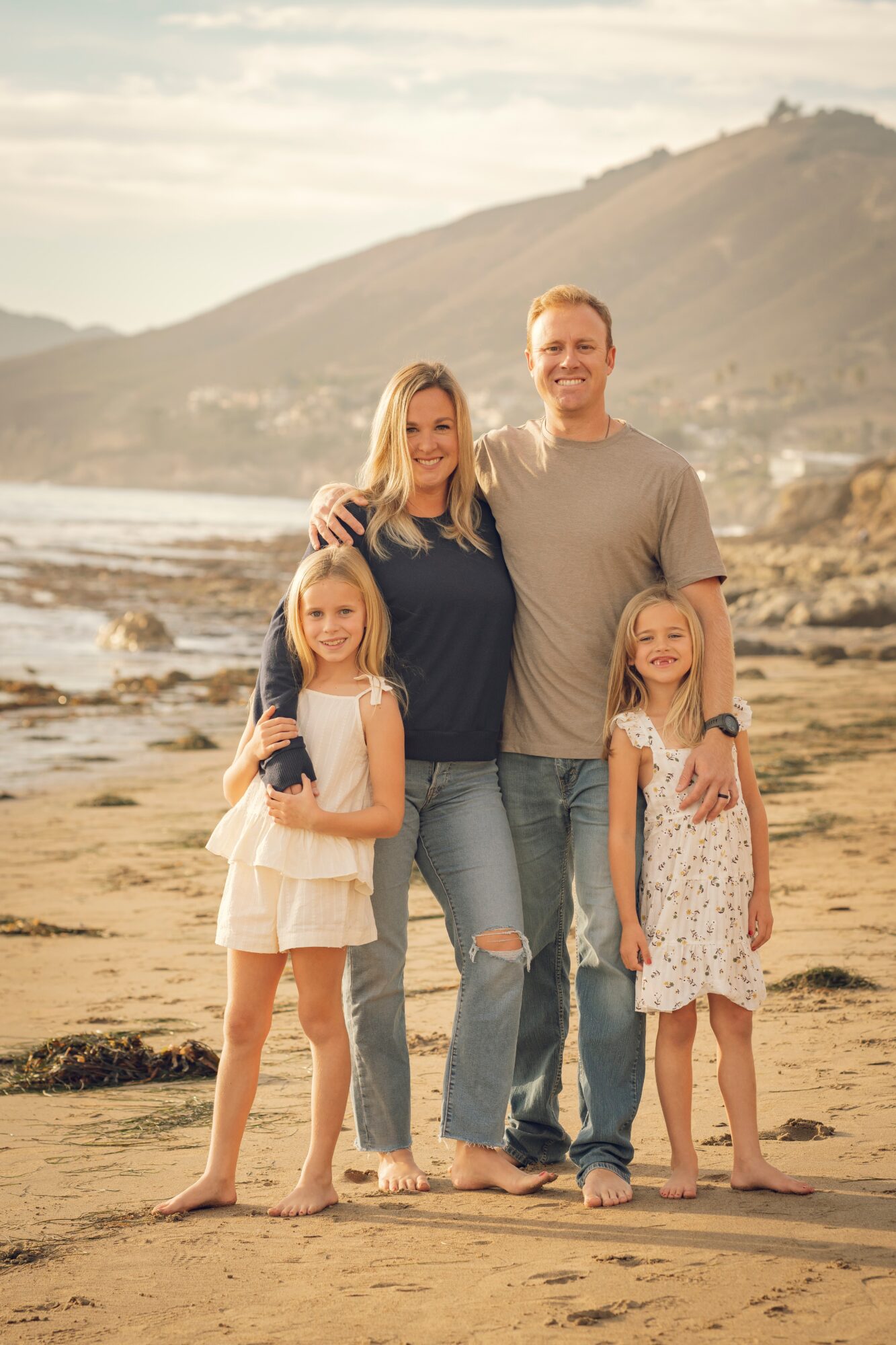 Family of four standing on a beach with hills in the background, smiling at the camera.