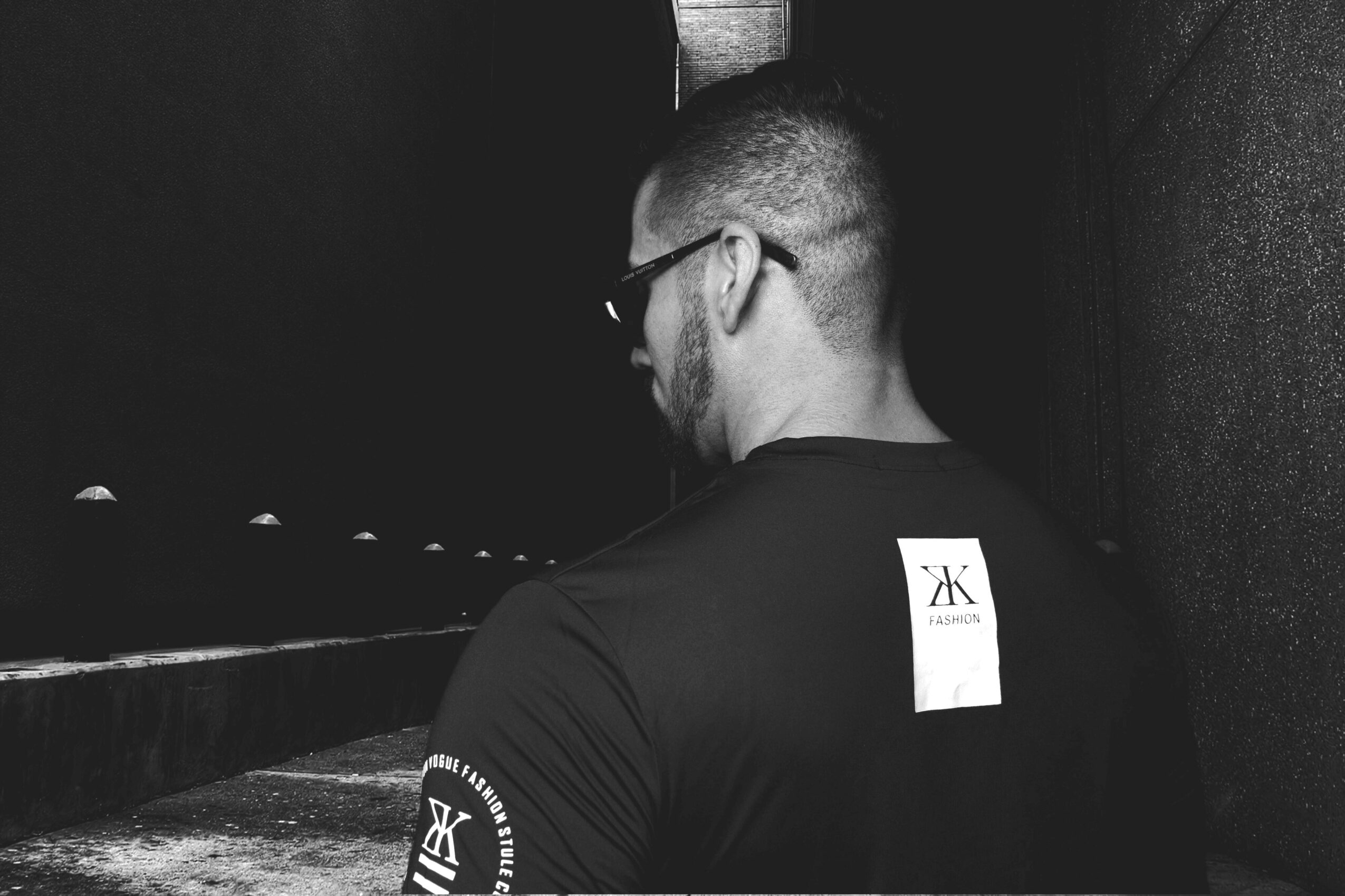 Man with glasses and a beard, facing away, standing in a dark space, wearing a black shirt with a logo on the back.