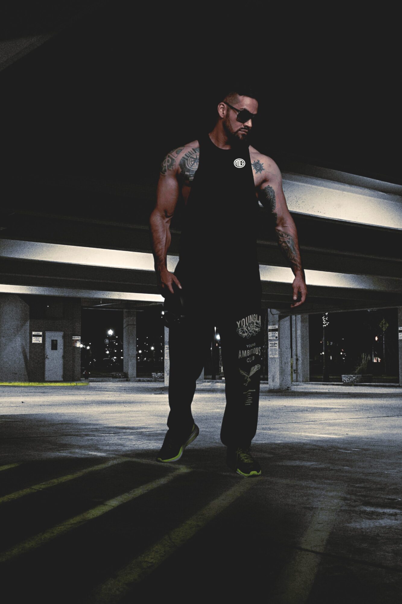 Man with tattoos and sunglasses walking in a parking garage, dimly lit, wearing black clothing.