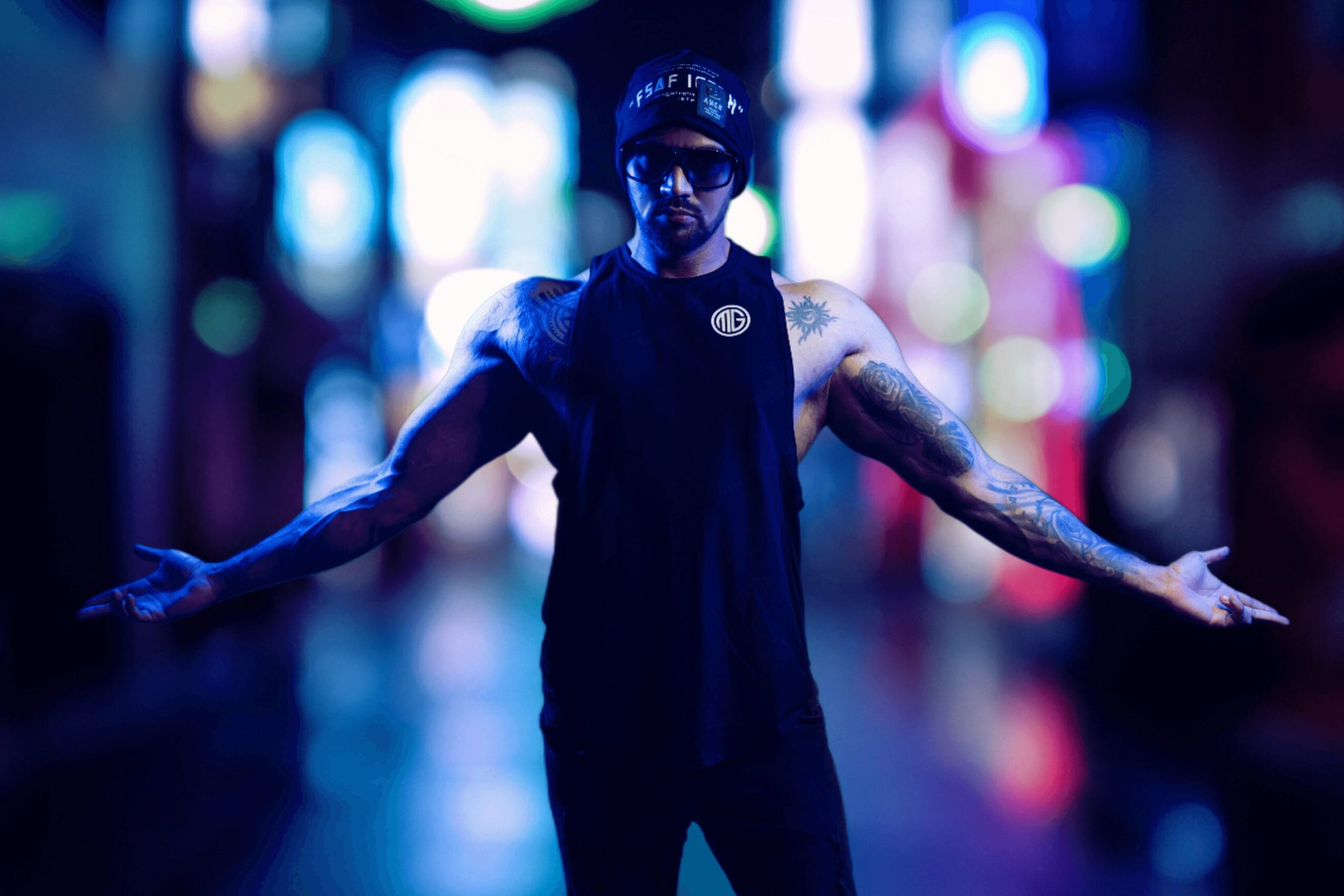 Person with arms outstretched standing in a colorful, blurred urban night scene, wearing dark clothing and sunglasses.