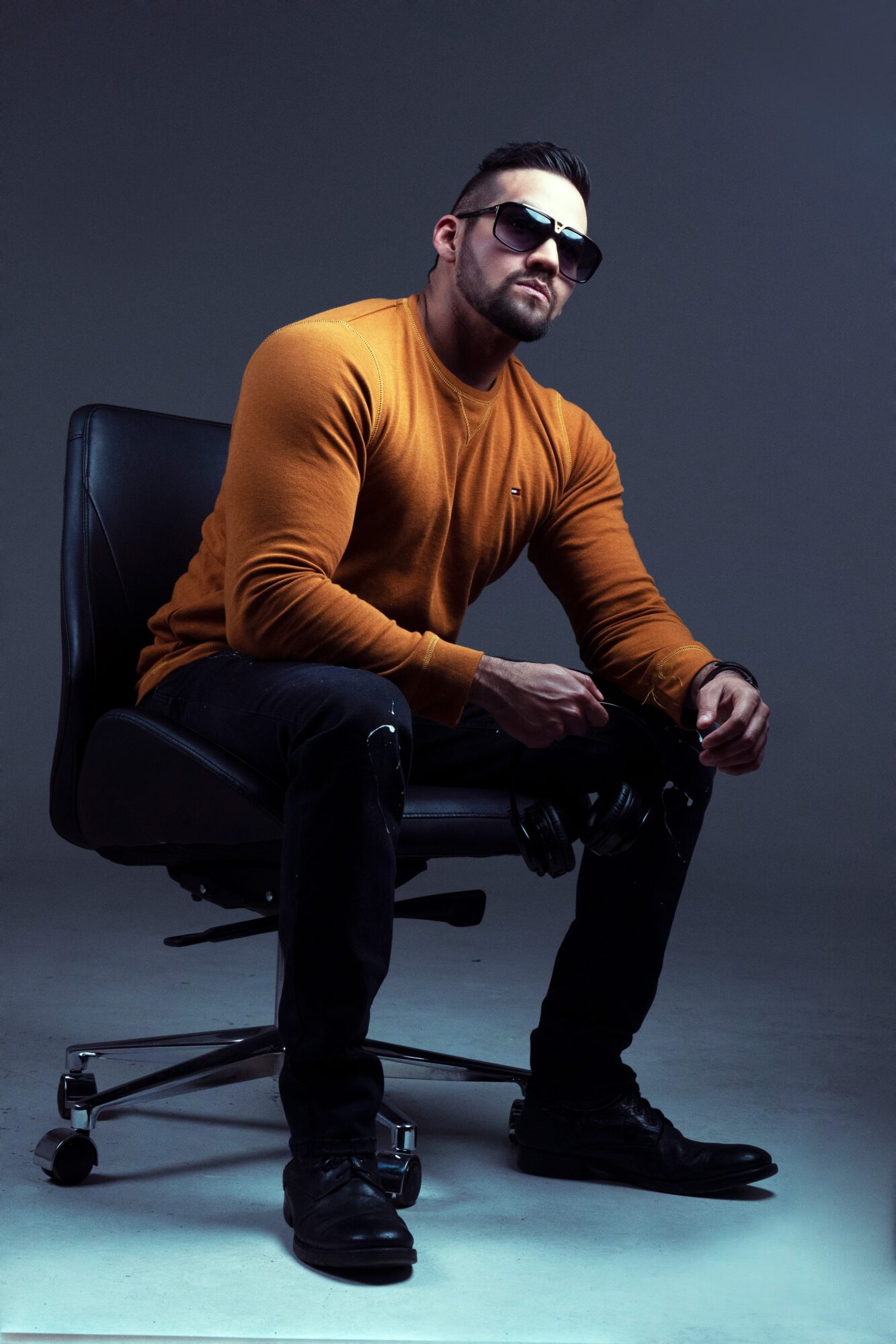 Man with sunglasses sitting on a chair, wearing an orange long-sleeve shirt and black pants, against a dark background.